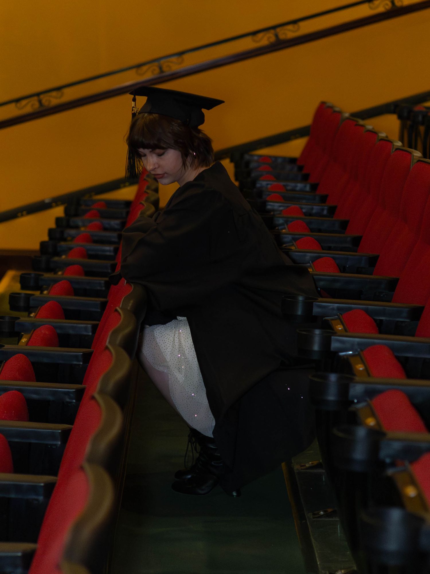 Graduate in cap and gown sitting on theater steps among red auditorium seats.