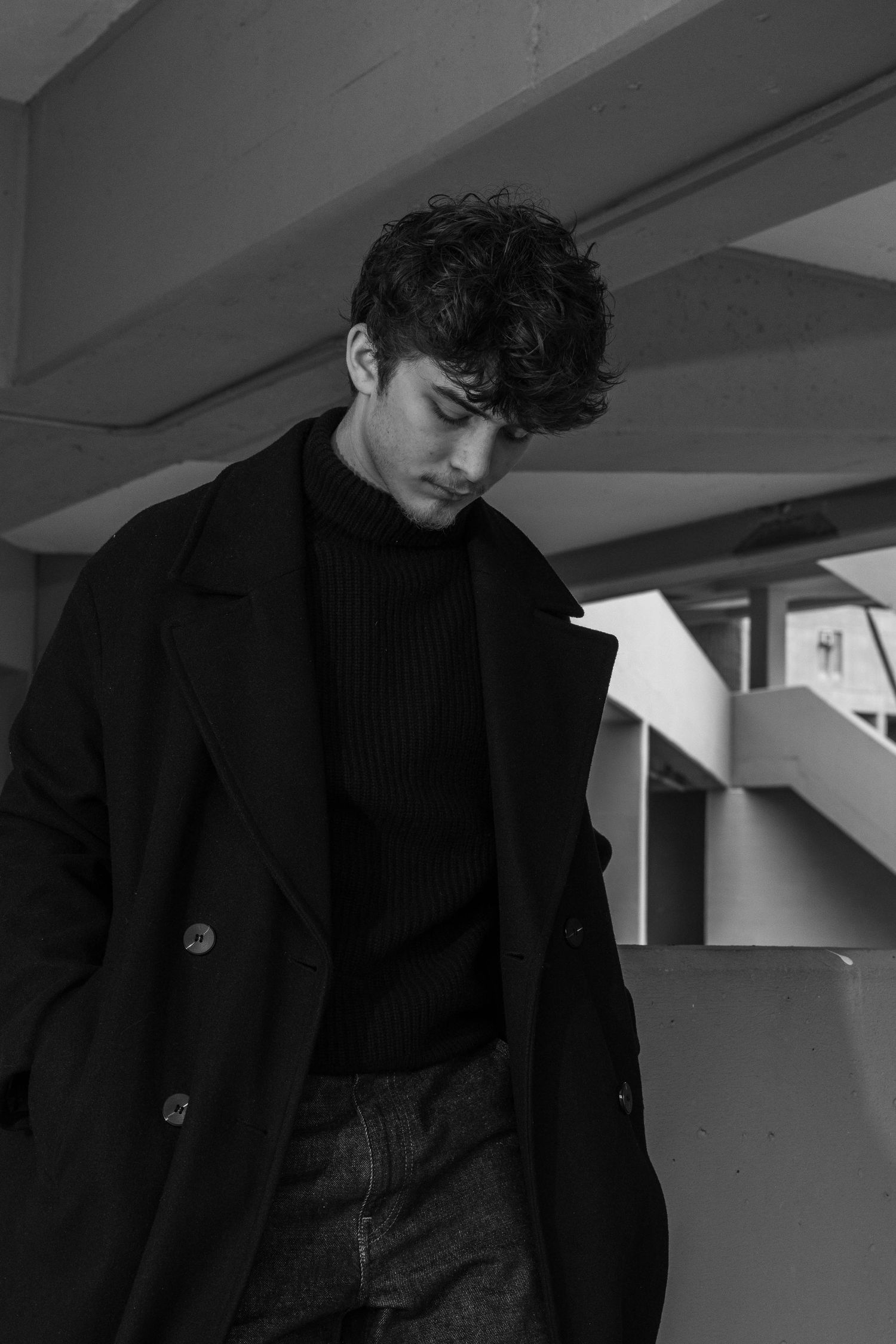 Black and white portrait of a young man with curly hair looking down, wearing a dark peacoat and turtleneck in a concrete parking structure.