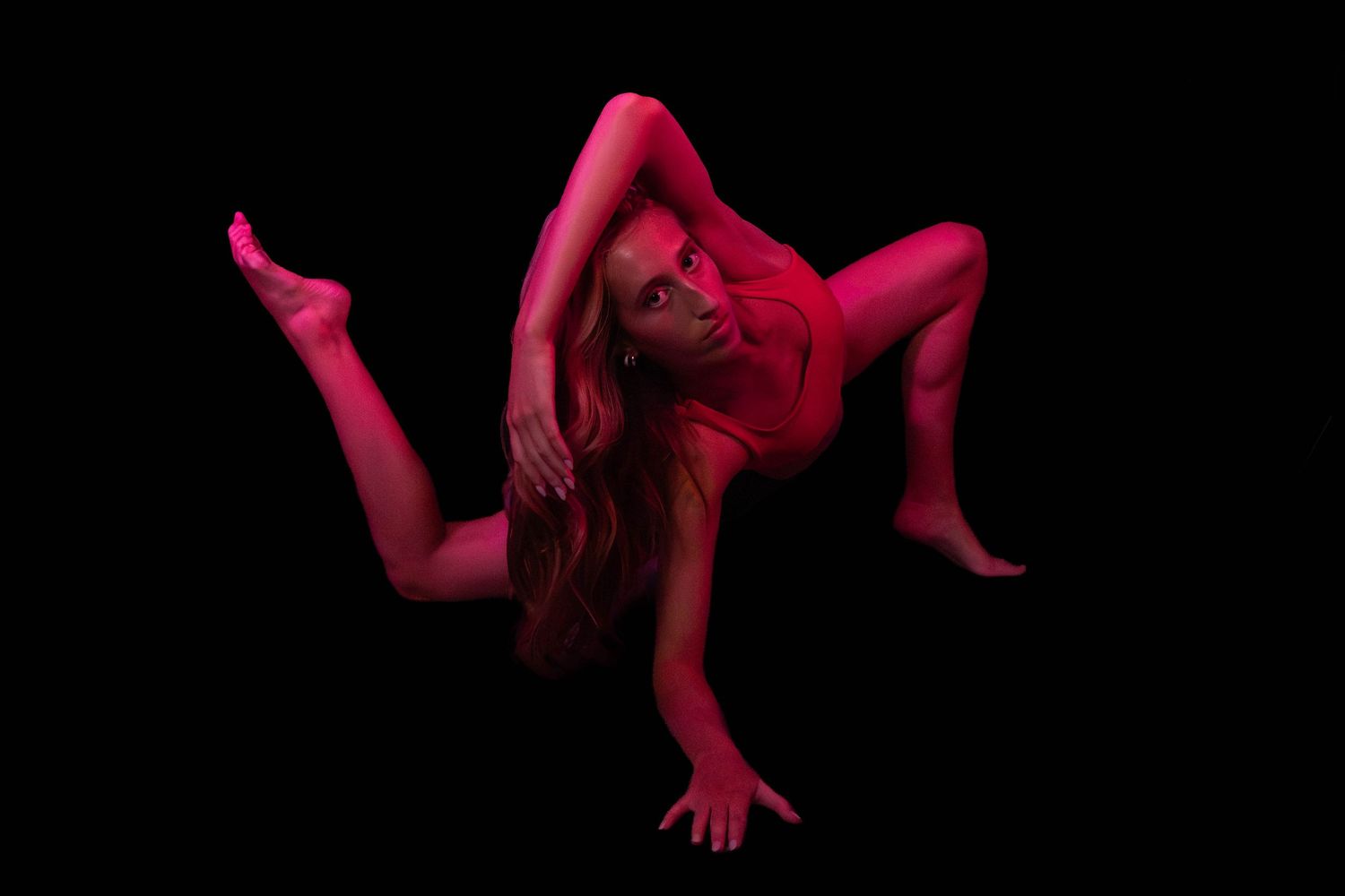 Contortionist in red-lit studio performing a deep backbend with legs splayed and arms braced against a black background.