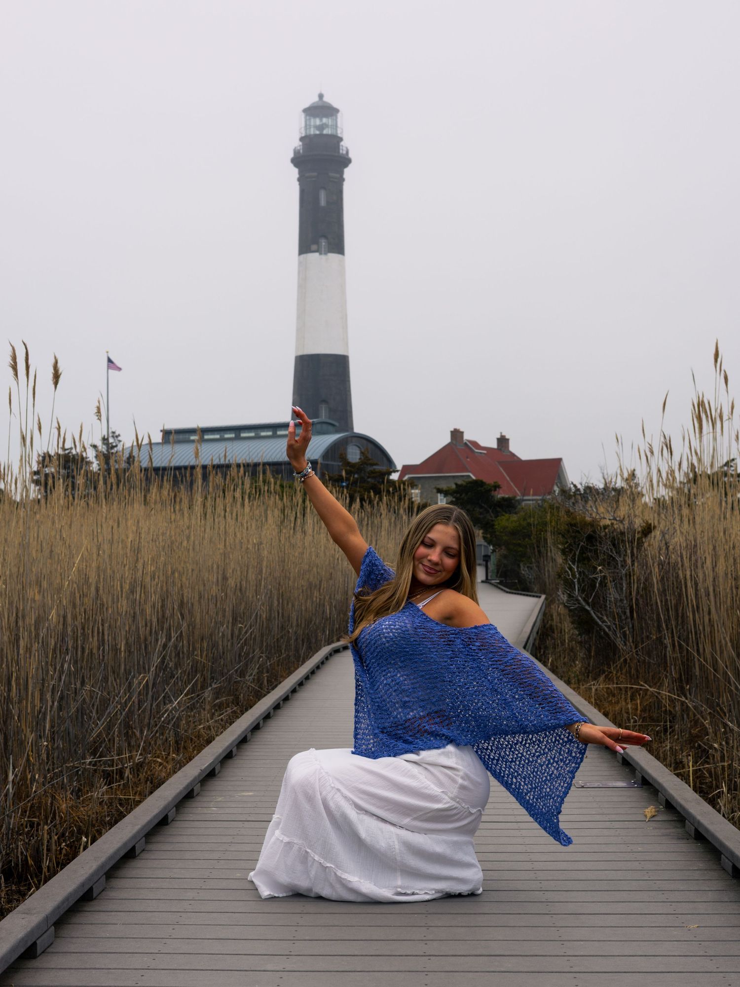Woman in a blue shawl and white skirt poses with raised arm on a wooden boardwalk with a black and white striped lighthouse behind her.