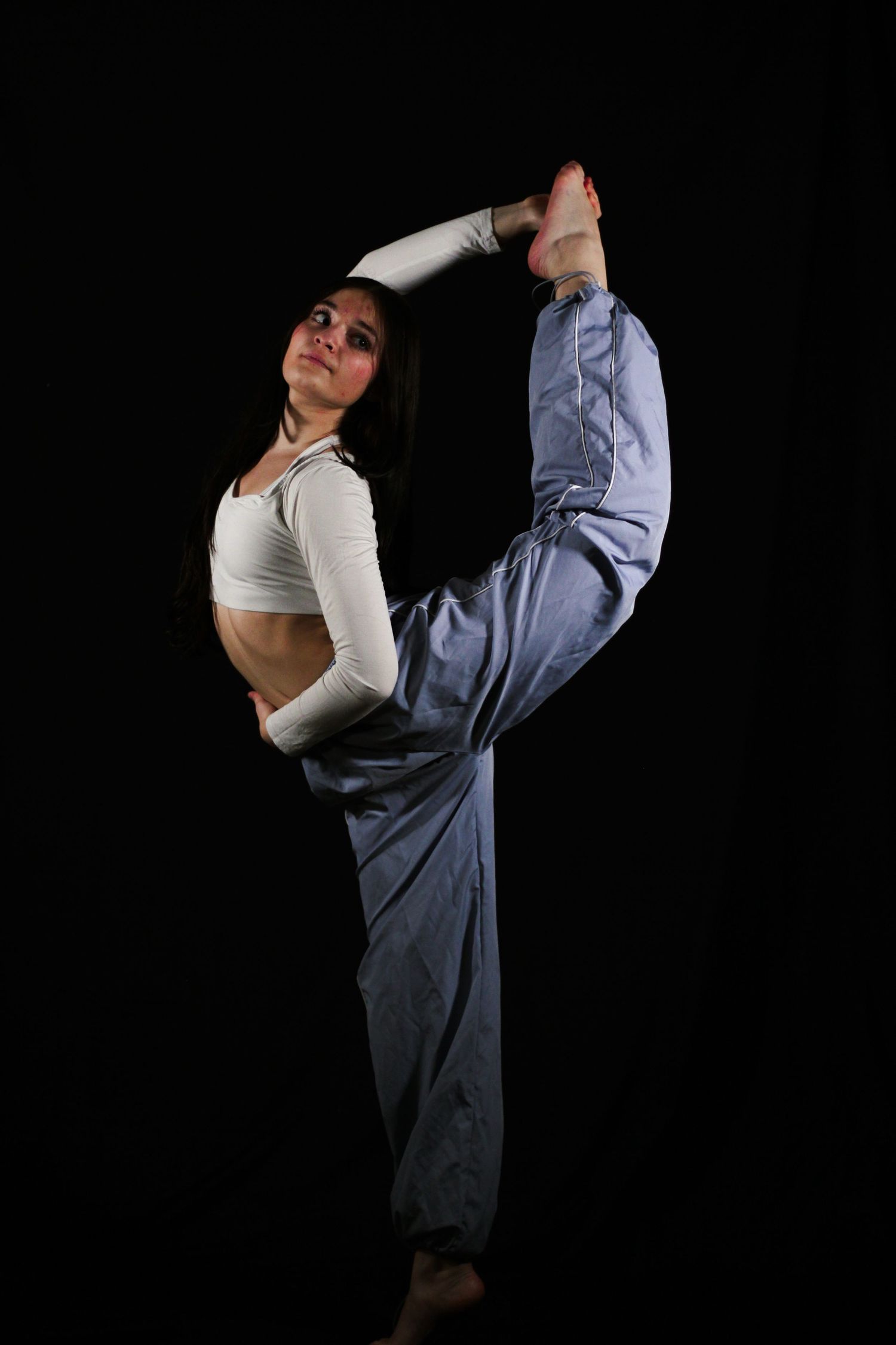 A flexible dancer in a white crop top and light blue pants performs a standing split against a dark background.