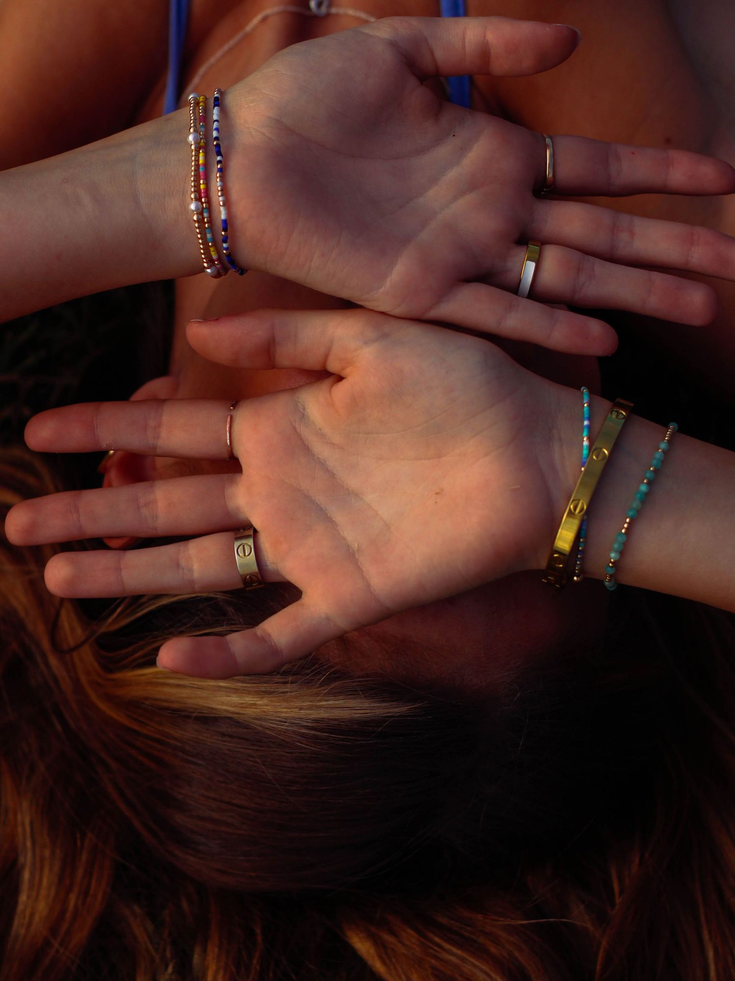Two hands adorned with colorful beaded bracelets and gold rings rest over a woman's face in warm golden light.