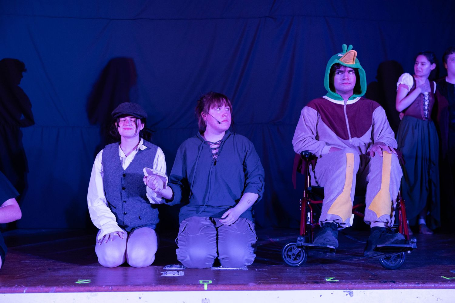 Performers in costumes kneel on stage during a theatrical production under colorful lights.