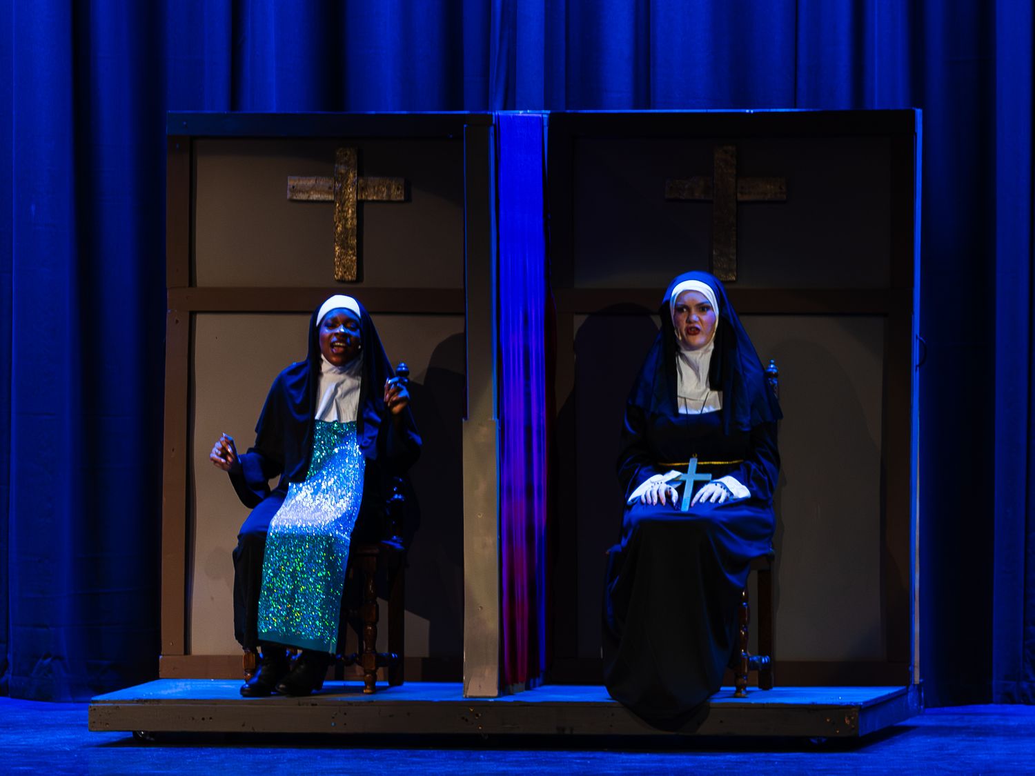 Two nuns sit in confessional booths on a blue-lit stage during a theatrical performance.
