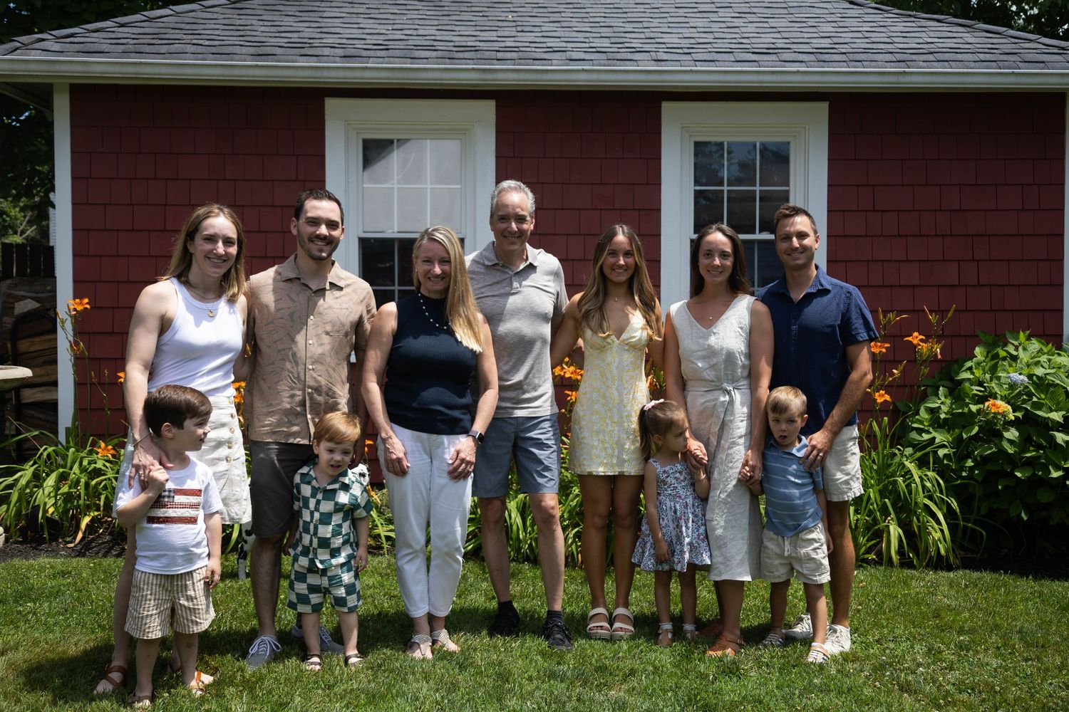 Large family group smiling together in front of a red cottage surrounded by summer gardens.
