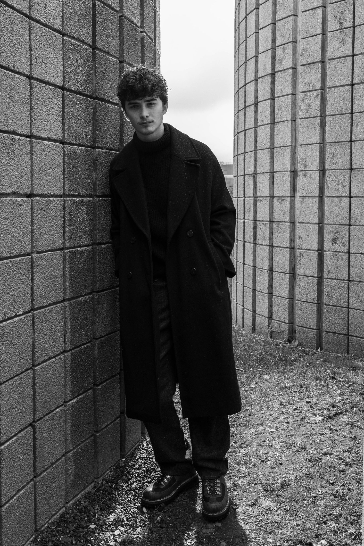 Black and white photo of a young man in a long dark overcoat and boots standing between two curved concrete block walls.