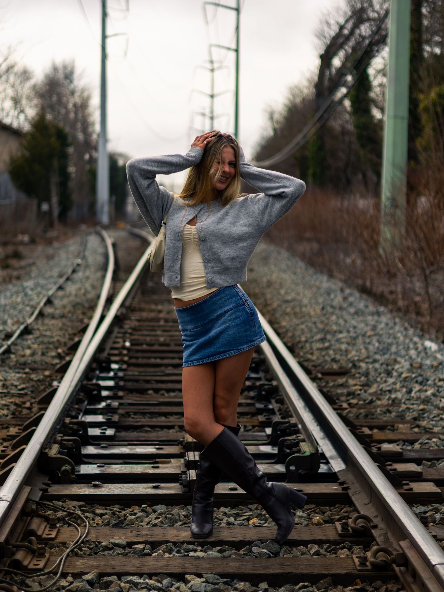 Young woman in a denim skirt, gray cardigan and black boots poses with a hand in her hair on railroad tracks under a cloudy sky.
