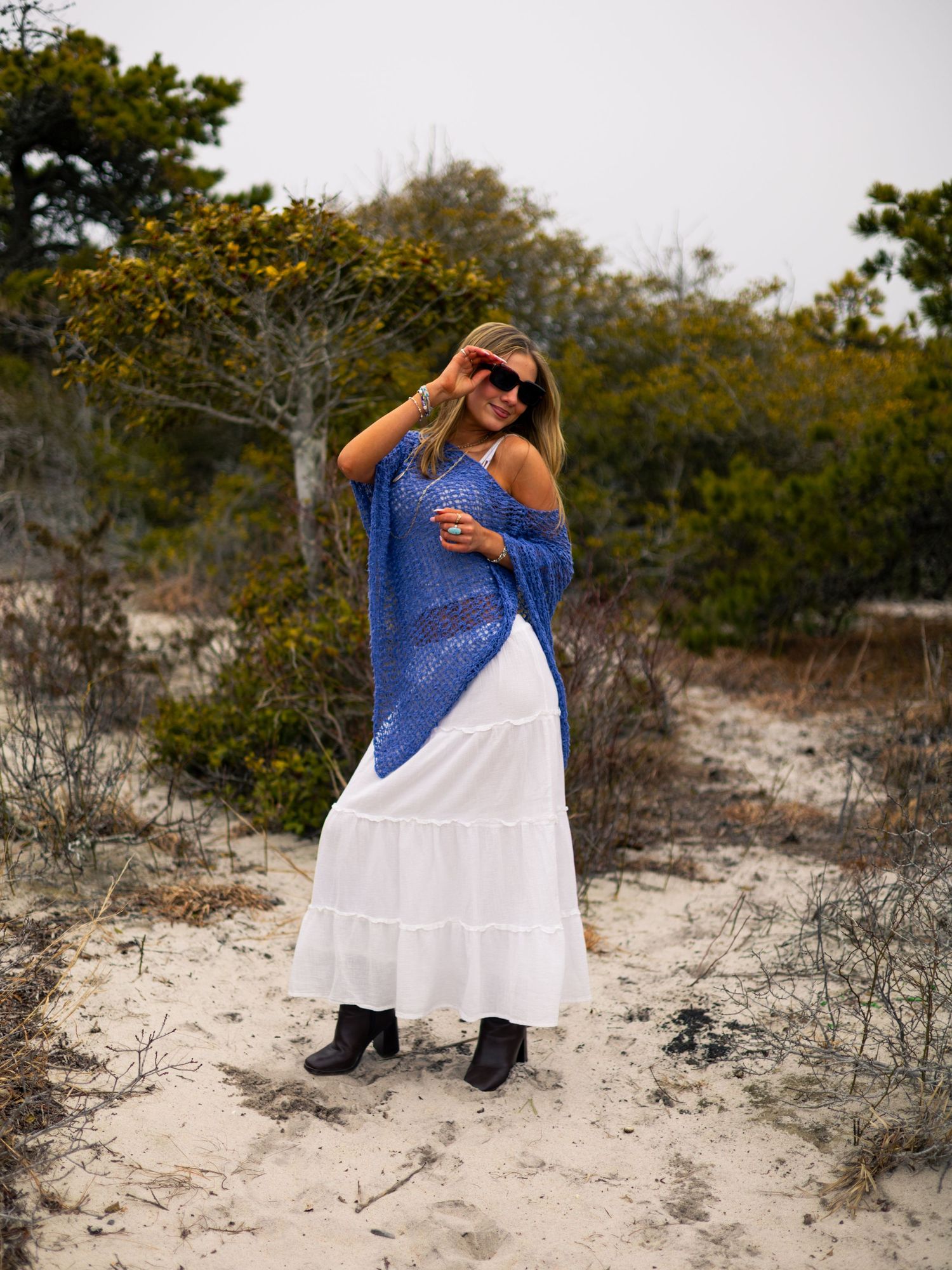 Woman in sunglasses wearing a blue crochet off-shoulder top, white tiered maxi skirt and black boots standing on a sandy coastal path.