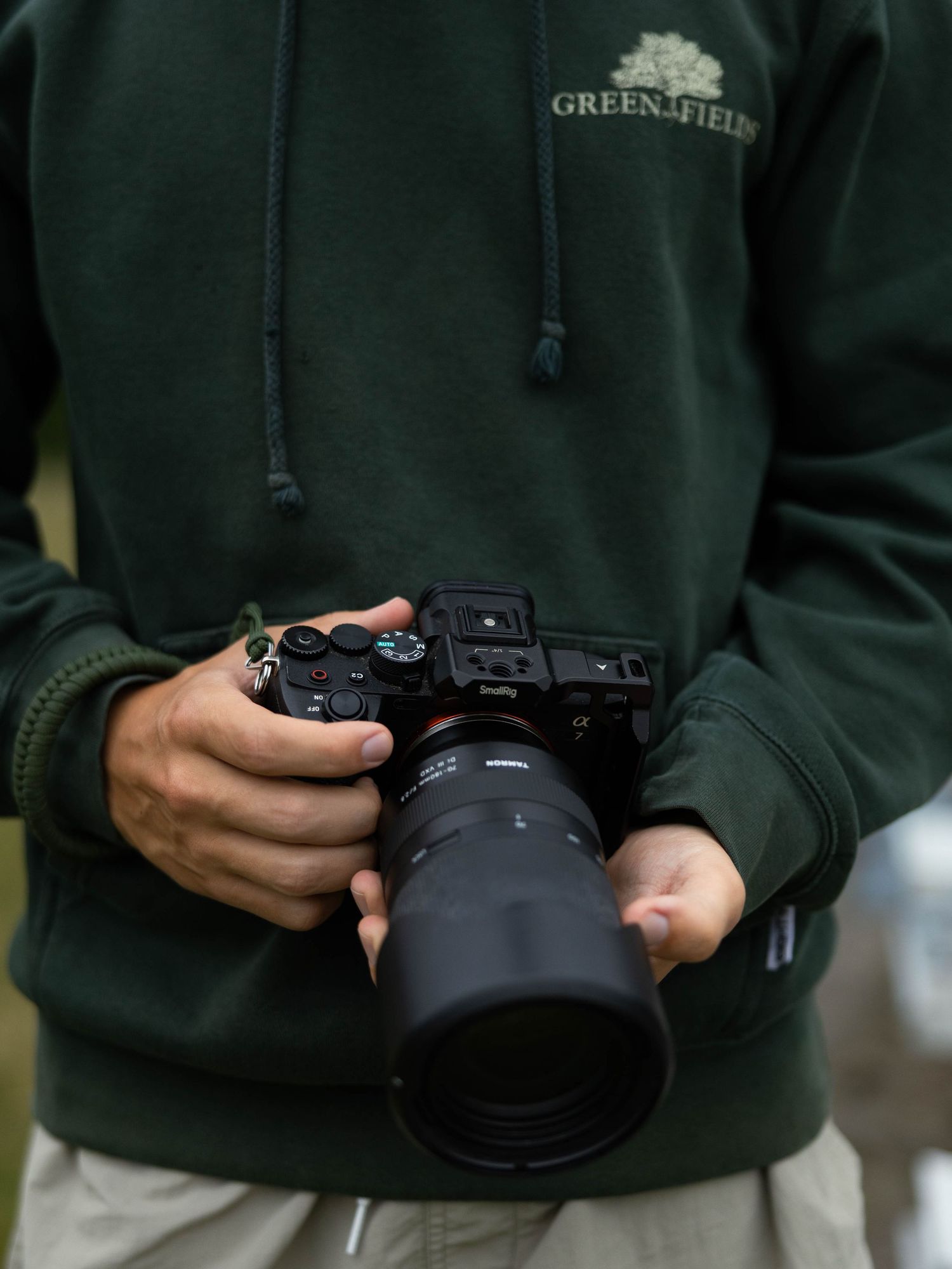 Person in green Greenfields hoodie holding a professional mirrorless camera with telephoto lens outdoors.