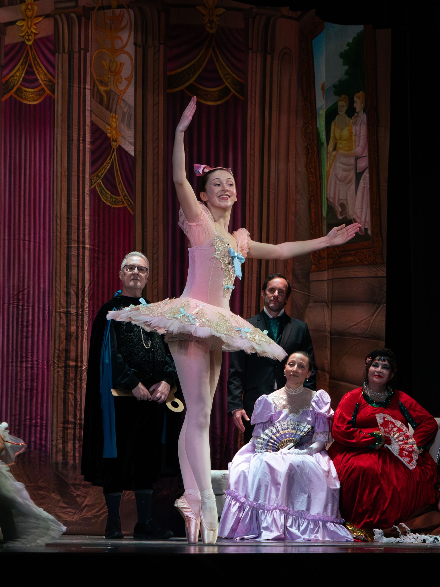 A ballerina in a pink tutu performs on stage en pointe, surrounded by costumed performers in an elegant theater setting.