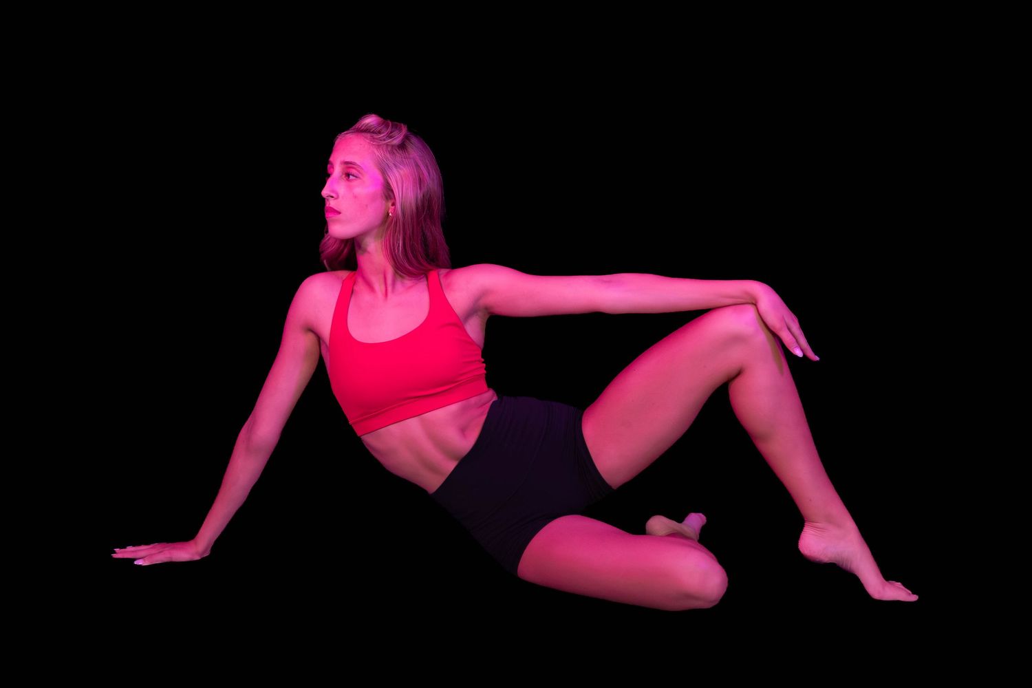 Female dancer in a red sports top and black shorts poses seated on the floor, lit by pink and red stage light against a black background.