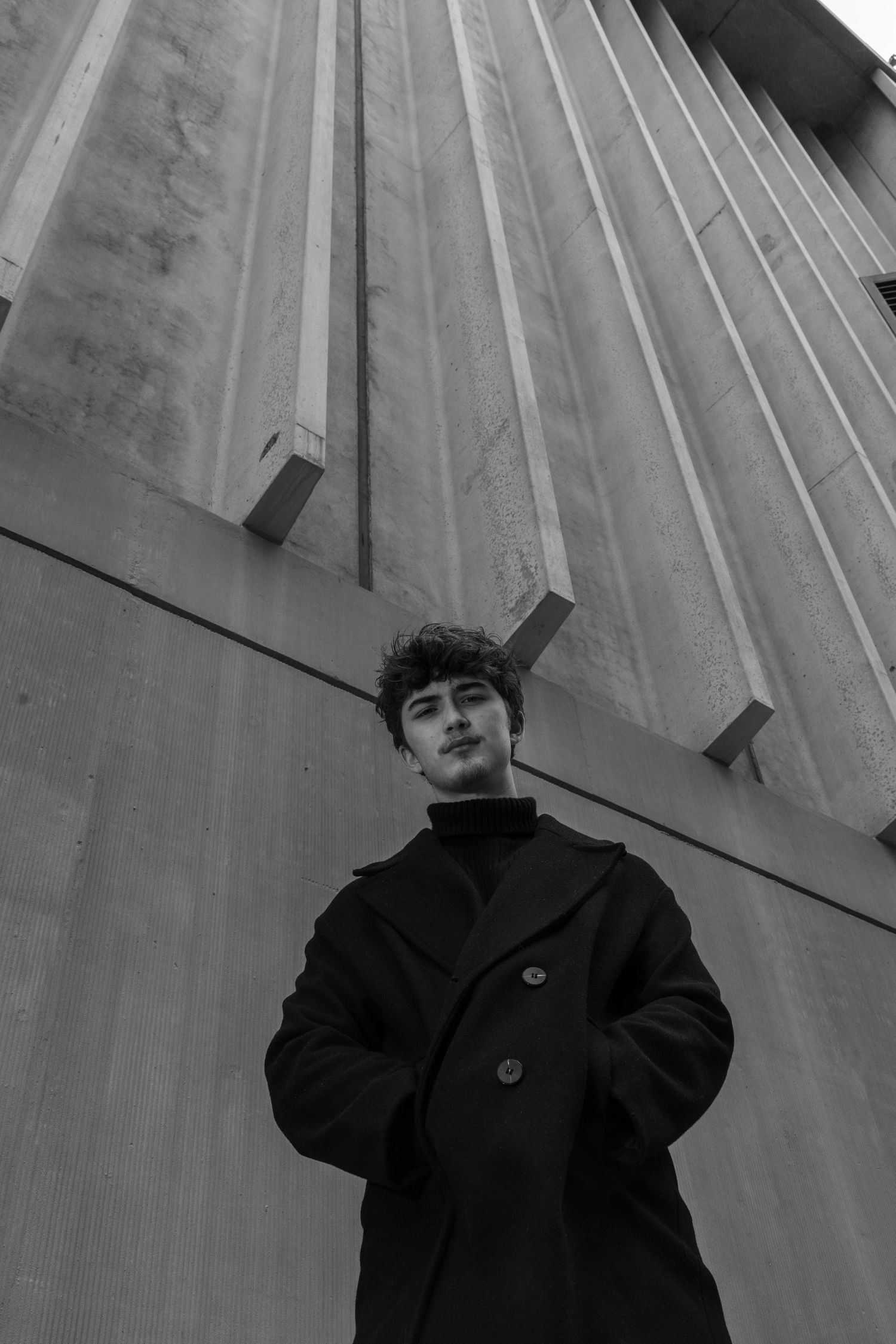 Black and white low-angle portrait of a curly-haired young man in a dark wool coat standing before a brutalist concrete building.