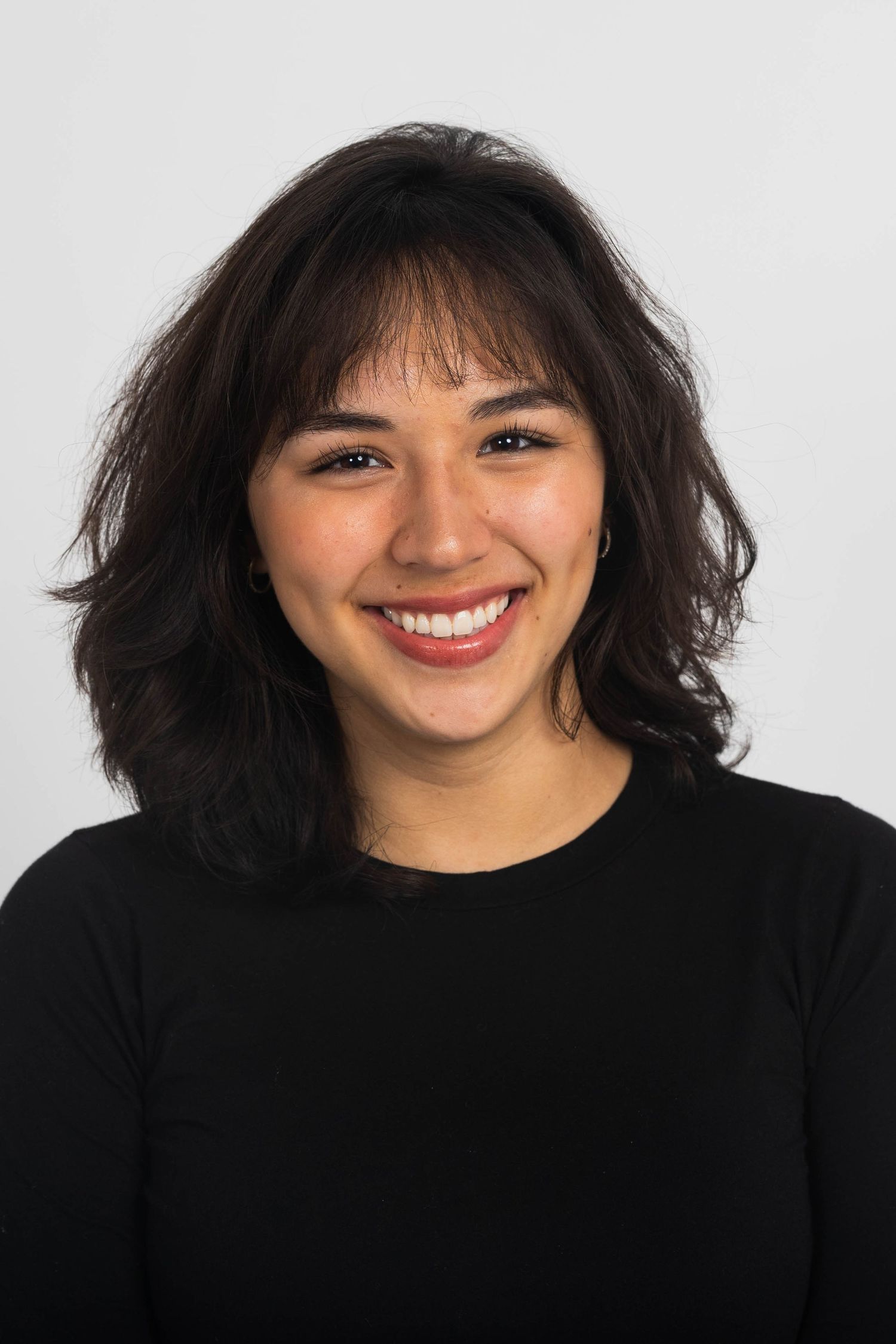 Smiling woman with shoulder-length dark wavy hair and bangs wearing a black top, posed against a plain light gray studio background.