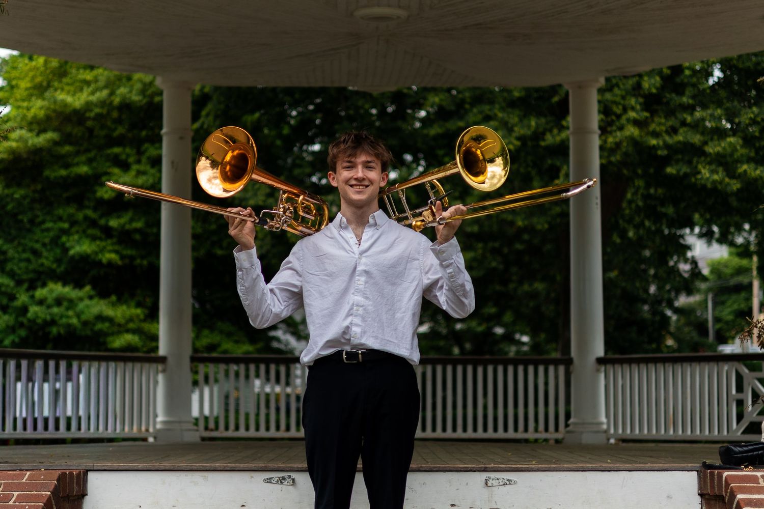 Young musician holding two trombones outdoors near a bandstand gazebo, wearing white shirt and black pants.