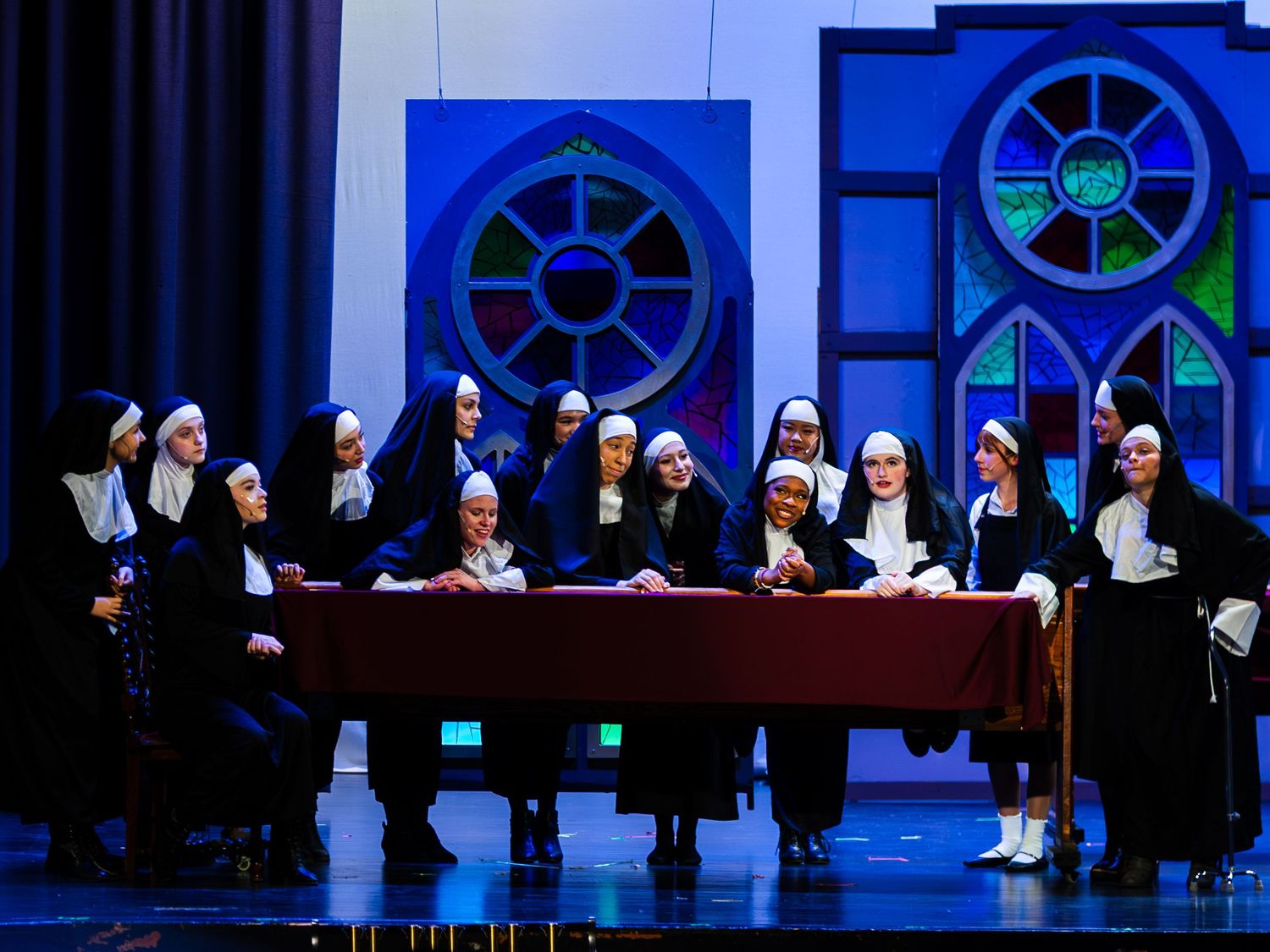 A large cast dressed as nuns performs a scene from Sister Act on a colorful church-themed stage.