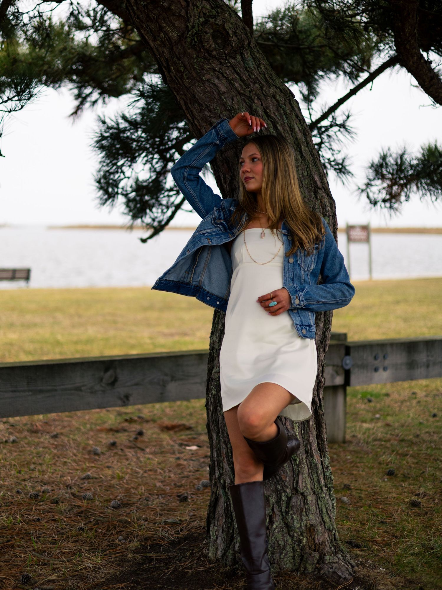 Young woman in white dress and denim jacket leaning against pine tree near waterfront on cloudy day.
