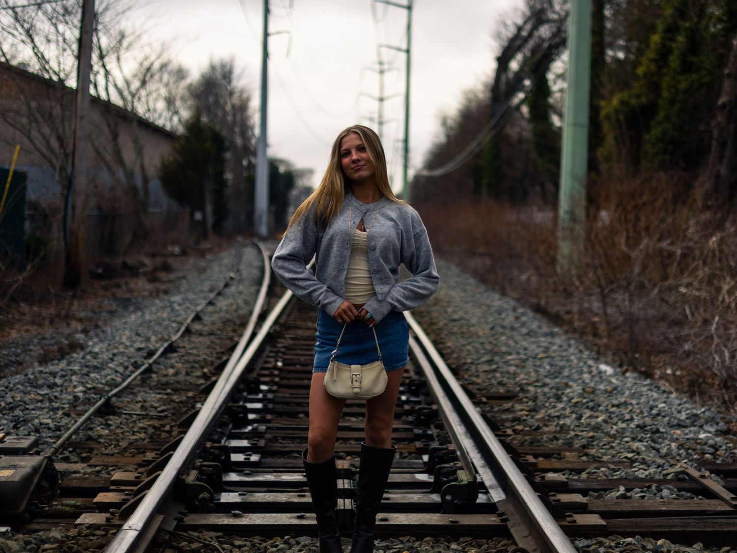 Blonde woman in denim jacket and mini skirt posing on railroad tracks surrounded by bare winter trees.