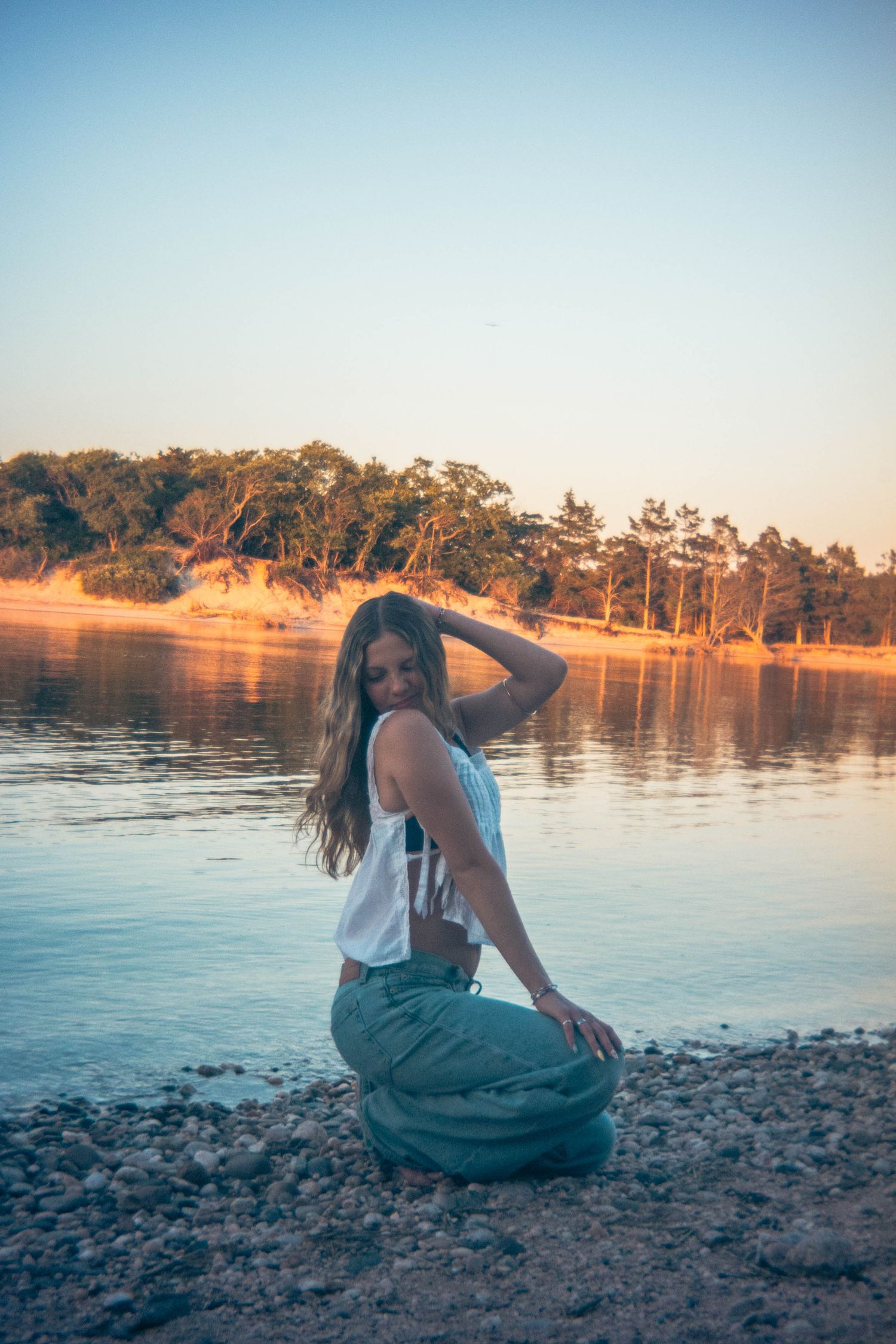 Smiling woman in white top and green maxi skirt kneeling on rocky shore at golden sunset.