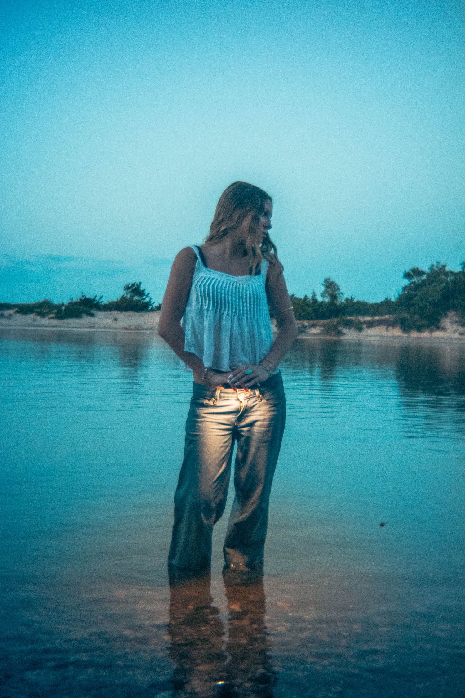Woman in white top and metallic pants stands in shallow water at dusk on a sandy shore.