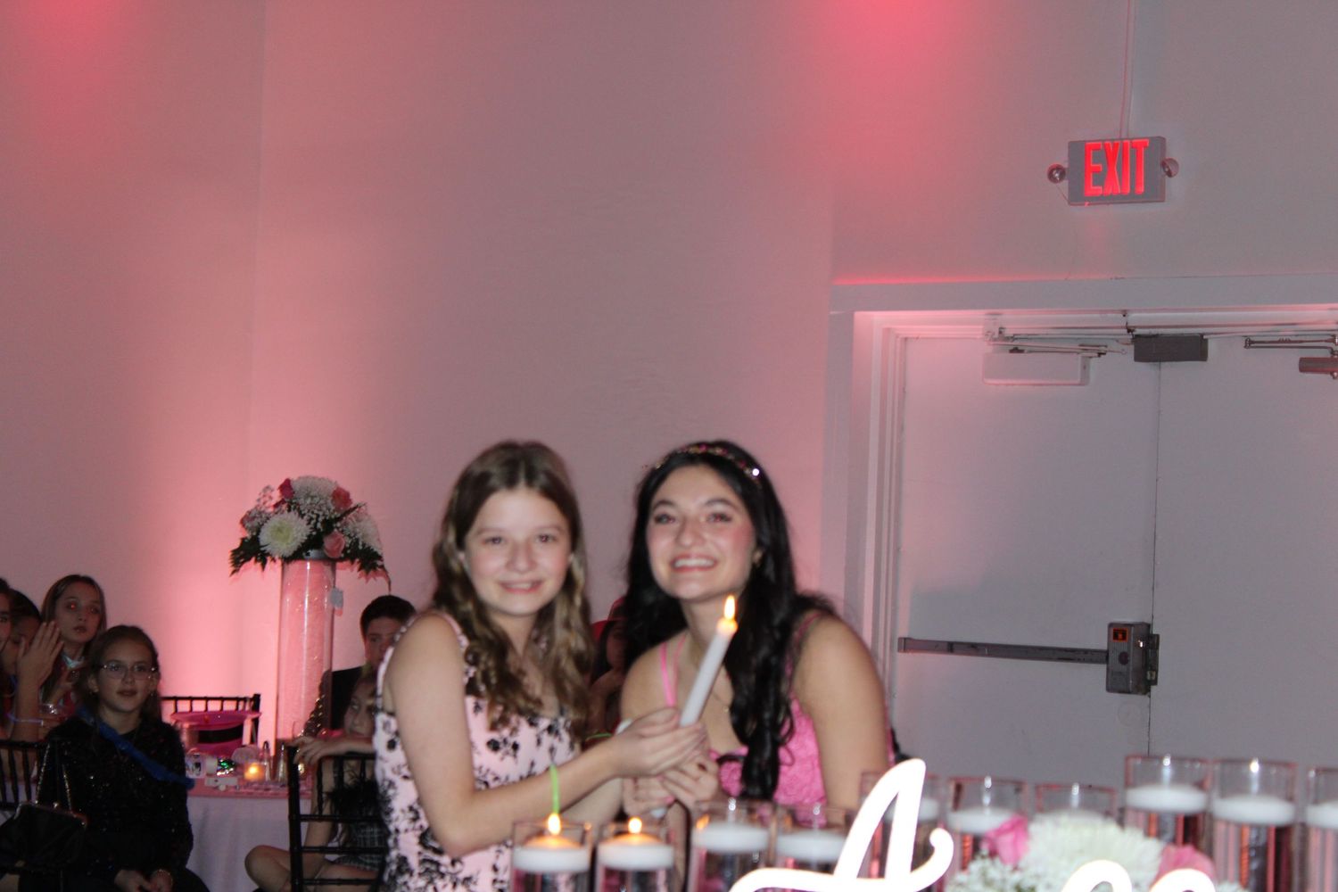 Two young girls smiling together at an elegant event with pink lighting and candles on the table.