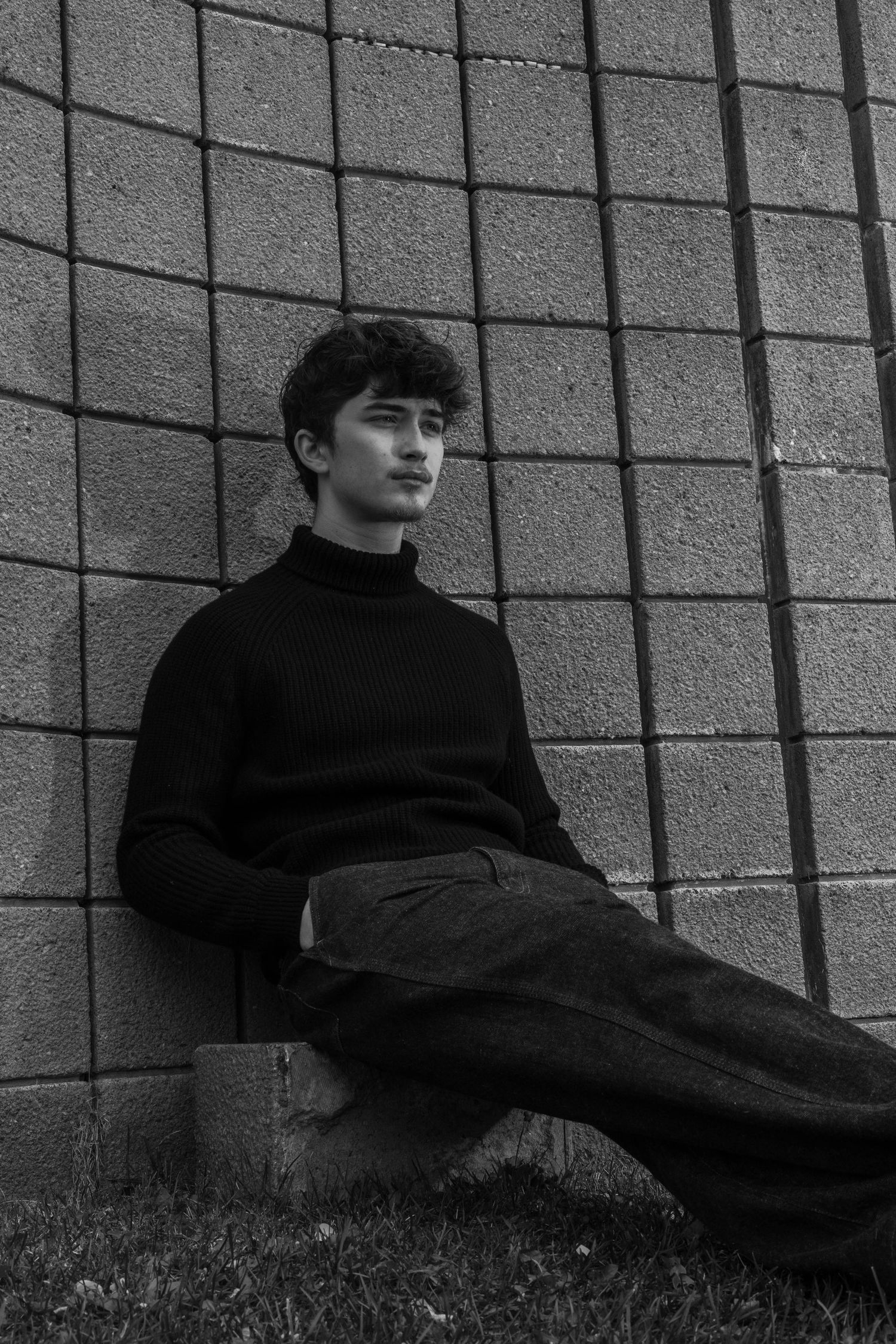 Black and white photo of a young man in a dark turtleneck and trousers sitting on a concrete block against a gray brick wall.