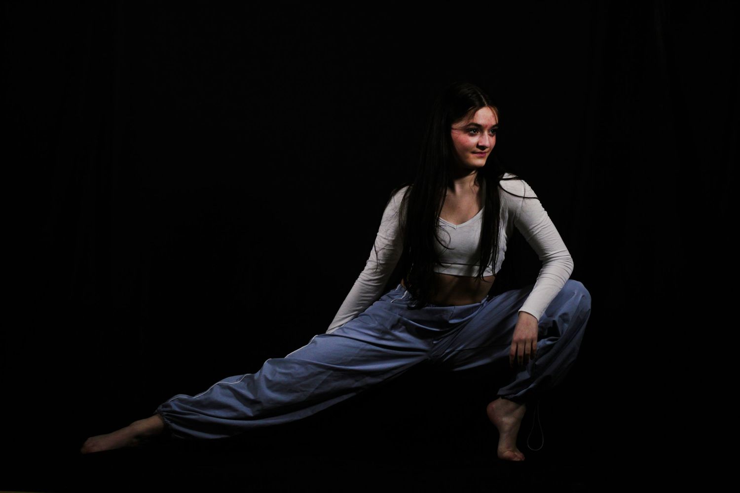 Young woman with long dark hair in a white top and blue baggy pants performs a deep side lunge stretch against a black studio background.