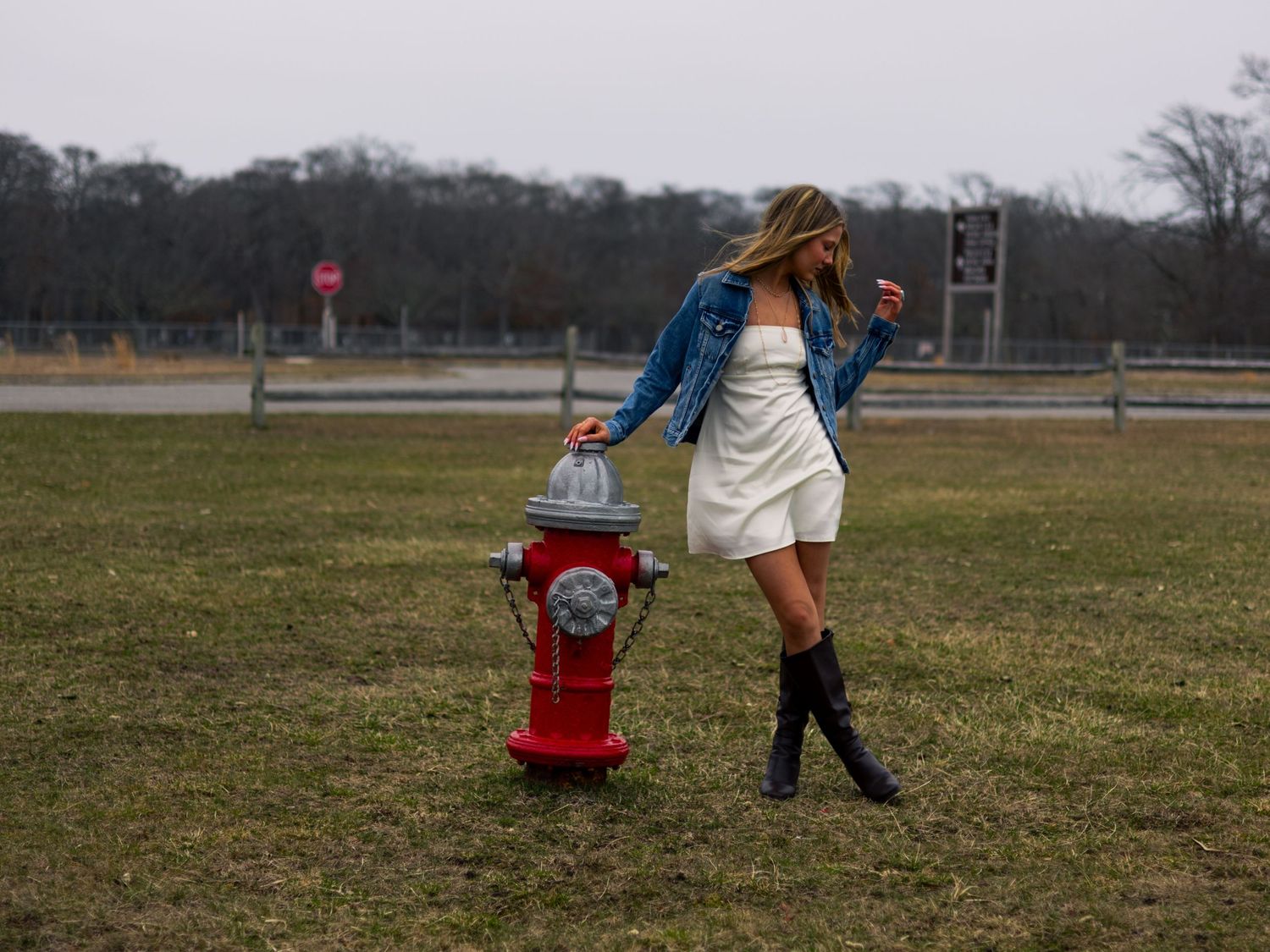 Woman in white dress and denim jacket posing next to a red fire hydrant in an open field.