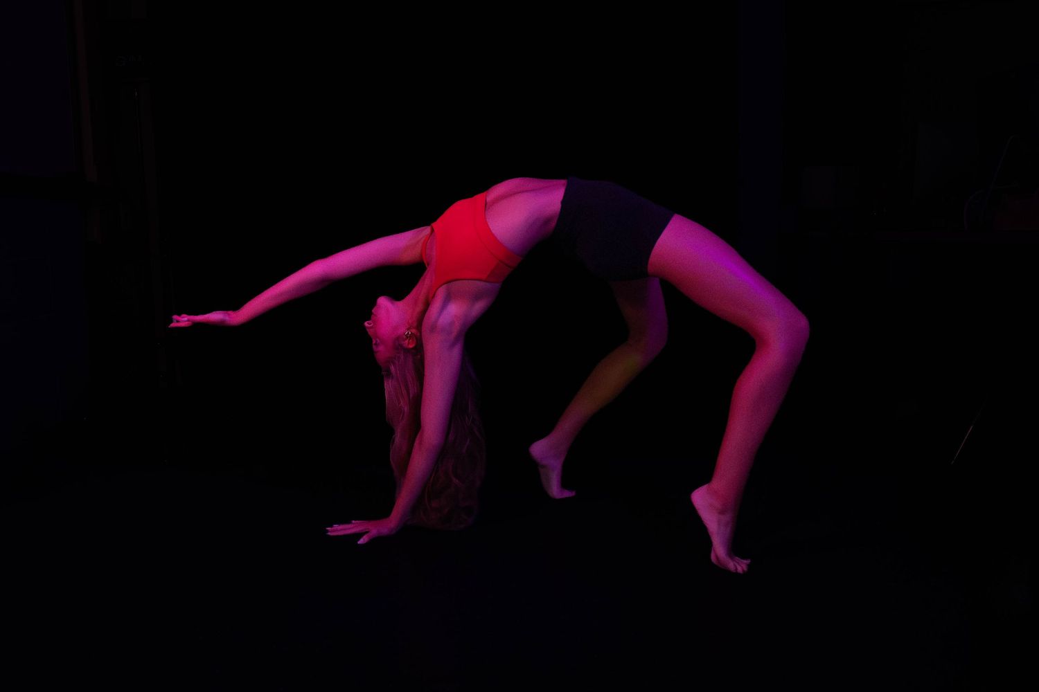 A contortionist in a red top and black shorts performs a deep backbend on a dark stage, lit by dramatic red lighting.