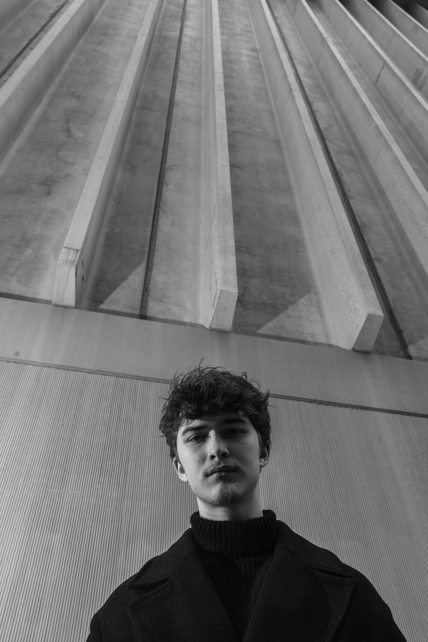 Black and white low-angle portrait of a young man with tousled hair in a dark coat and turtleneck beneath a ribbed concrete ceiling.