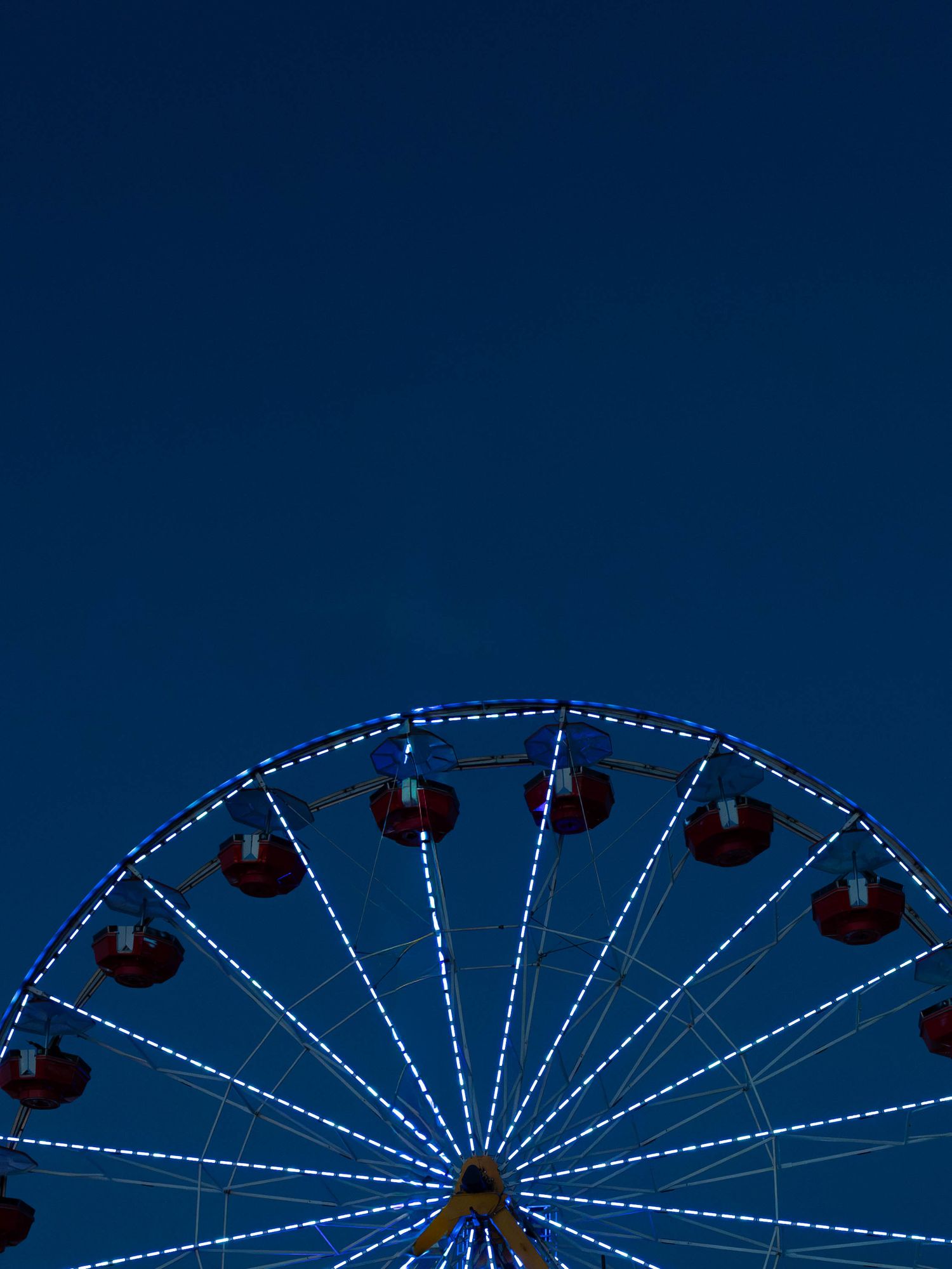 Illuminated ferris wheel with red gondolas and blue LED lights against a deep blue twilight sky at an amusement park.