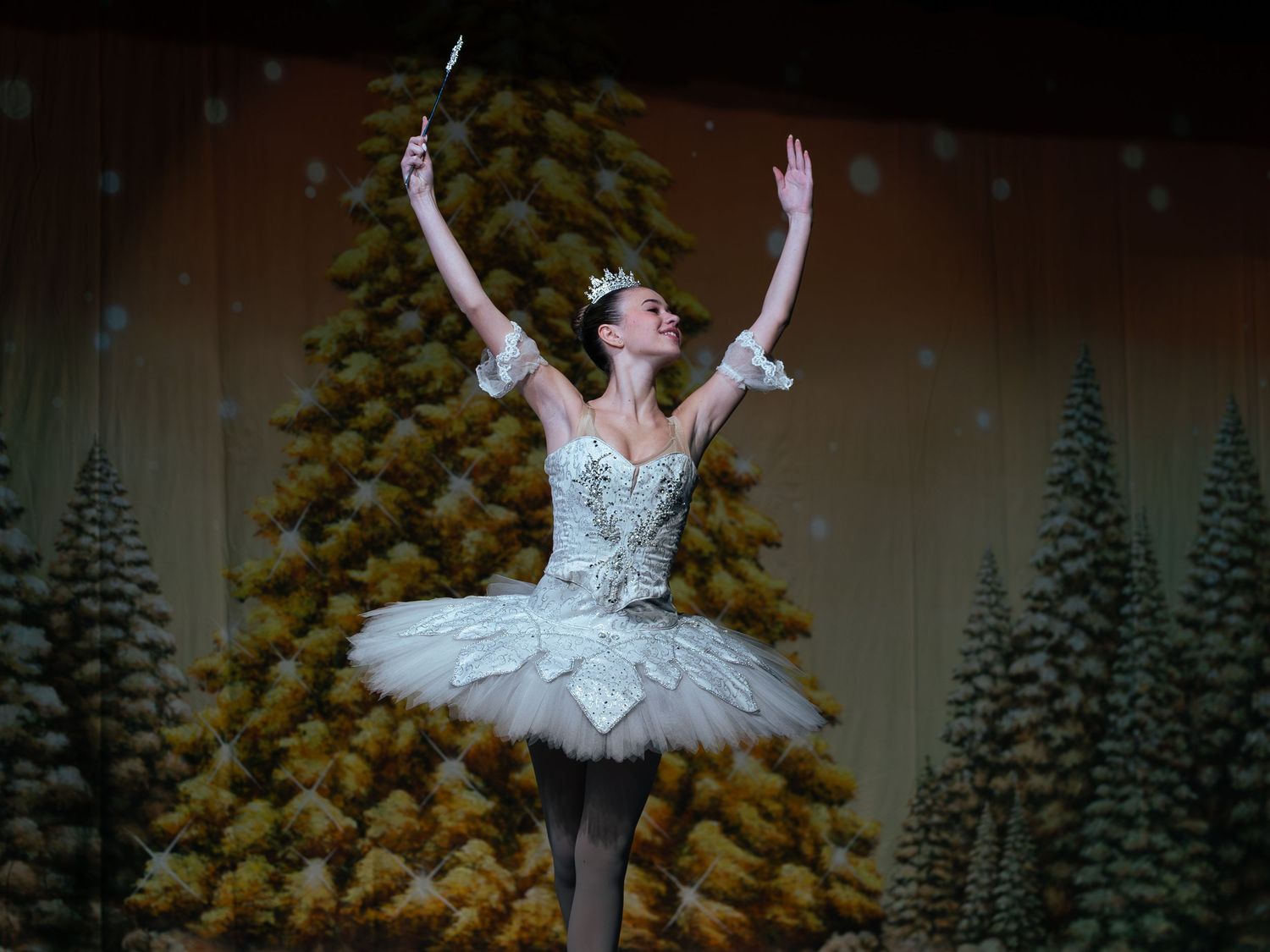 A ballerina in a white tutu and crown performs on stage before a Christmas tree backdrop in The Nutcracker ballet.