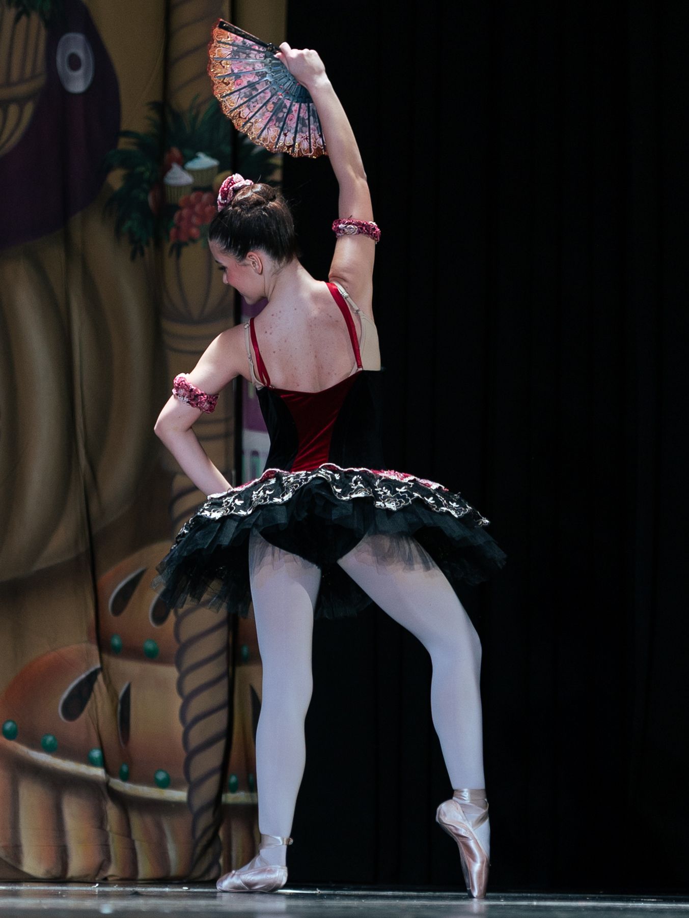 Ballerina in red and black tutu performs on pointe, holding a decorative fan overhead on stage.