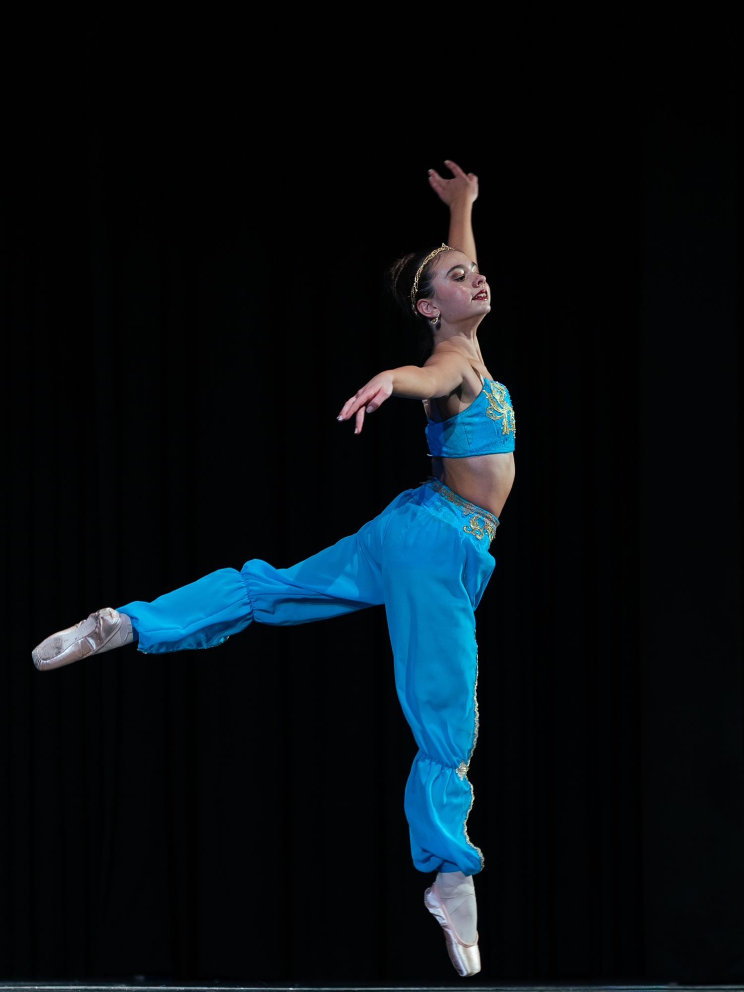 Ballet dancer in blue costume performs an elegant arabesque on pointe against a dark stage background.