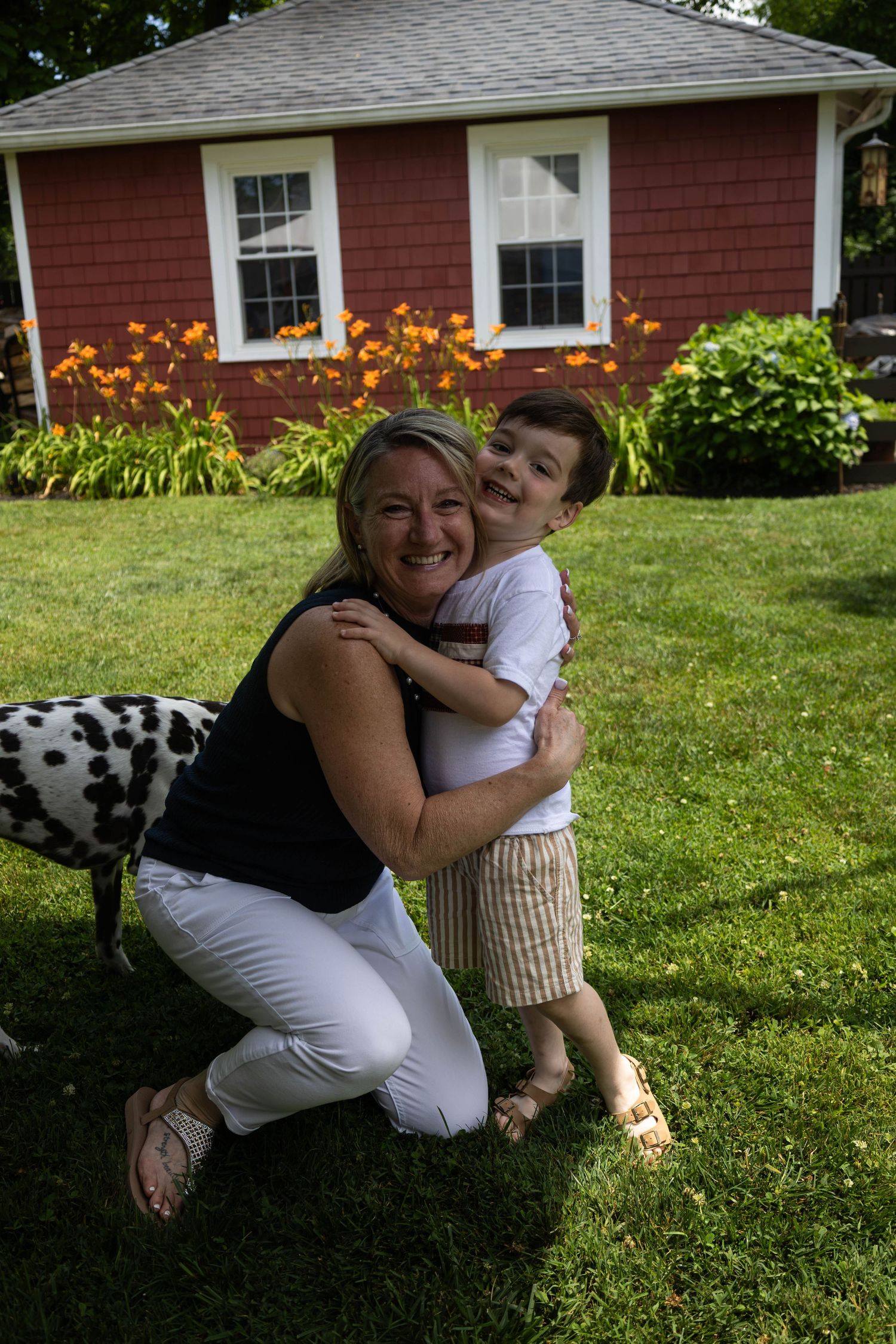 Smiling woman kneels on green lawn hugging a young boy with a Dalmatian dog beside a red house.
