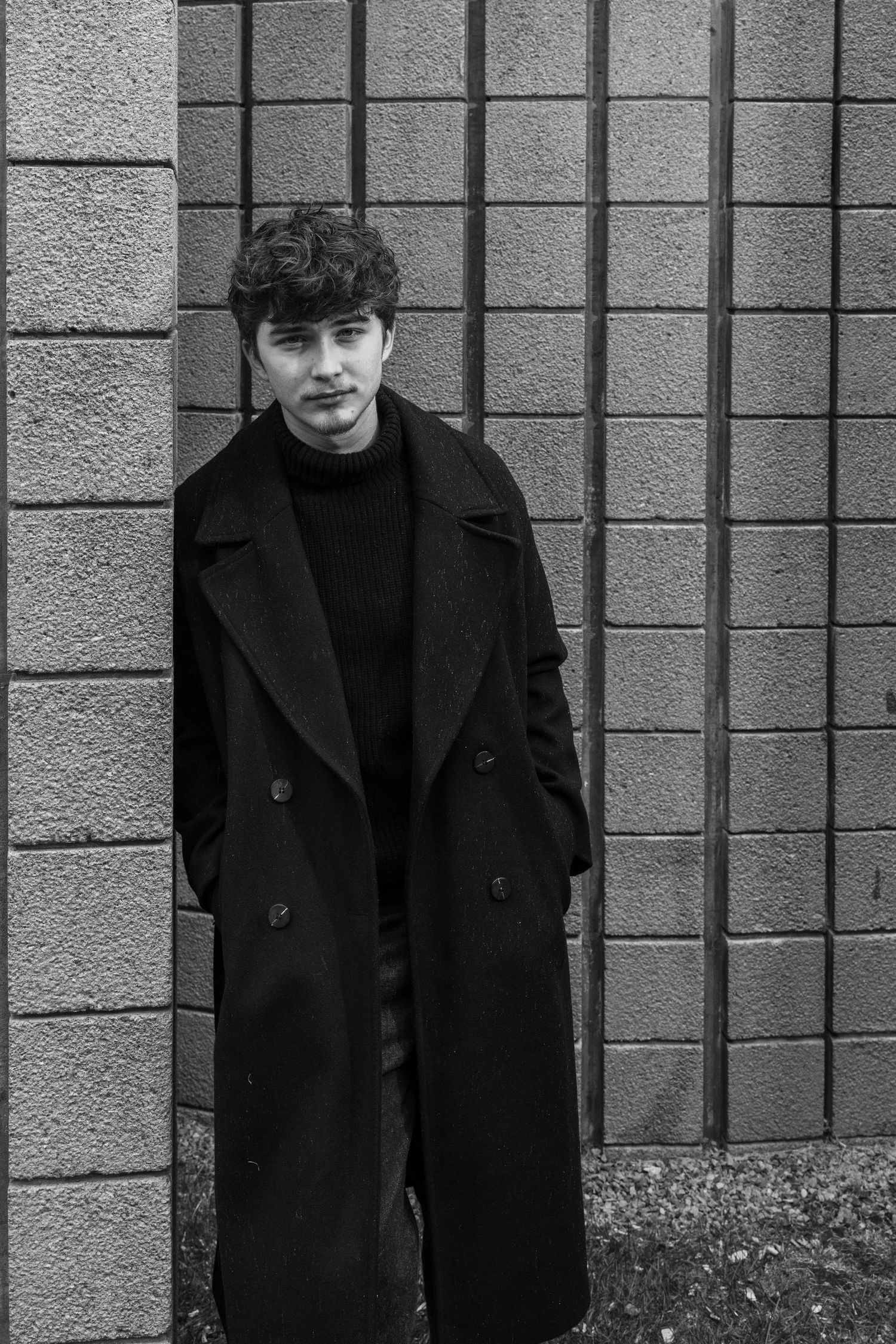 Black and white photo of a young man with curly hair in a long dark overcoat and turtleneck leaning against a concrete block wall.