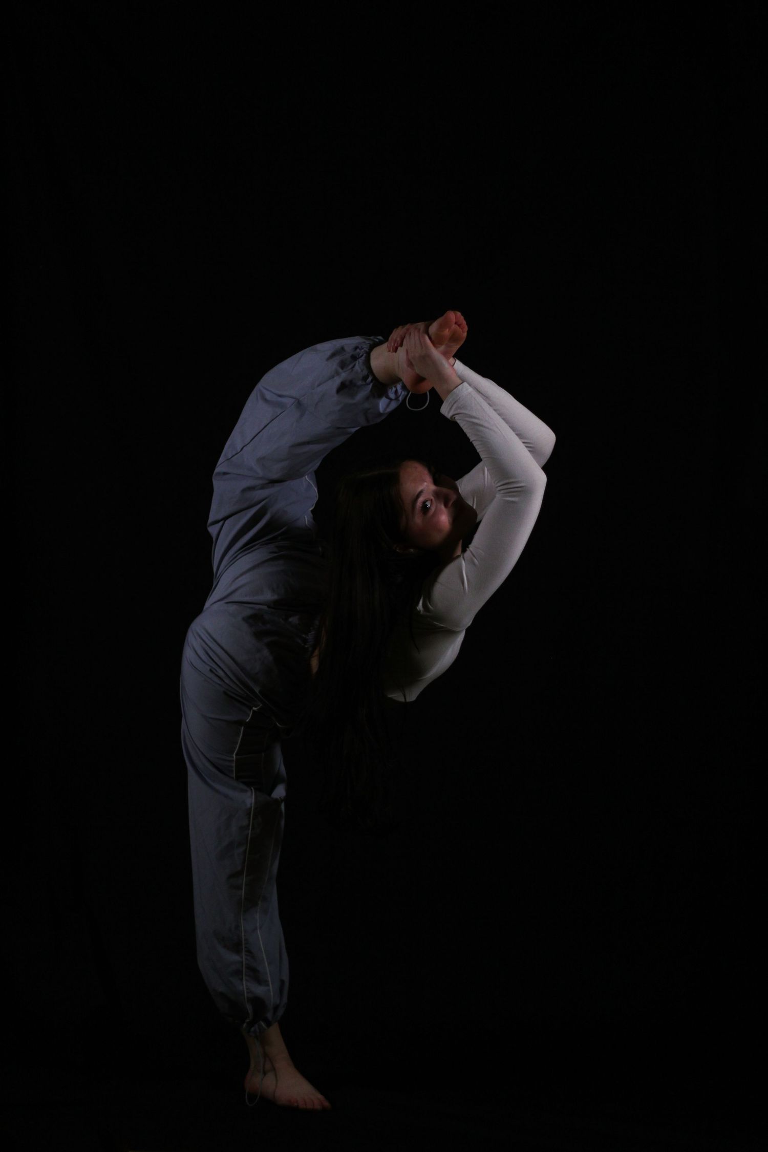 A flexible dancer in gray clothing performs a standing backbend, bending one leg overhead against a dark studio background.