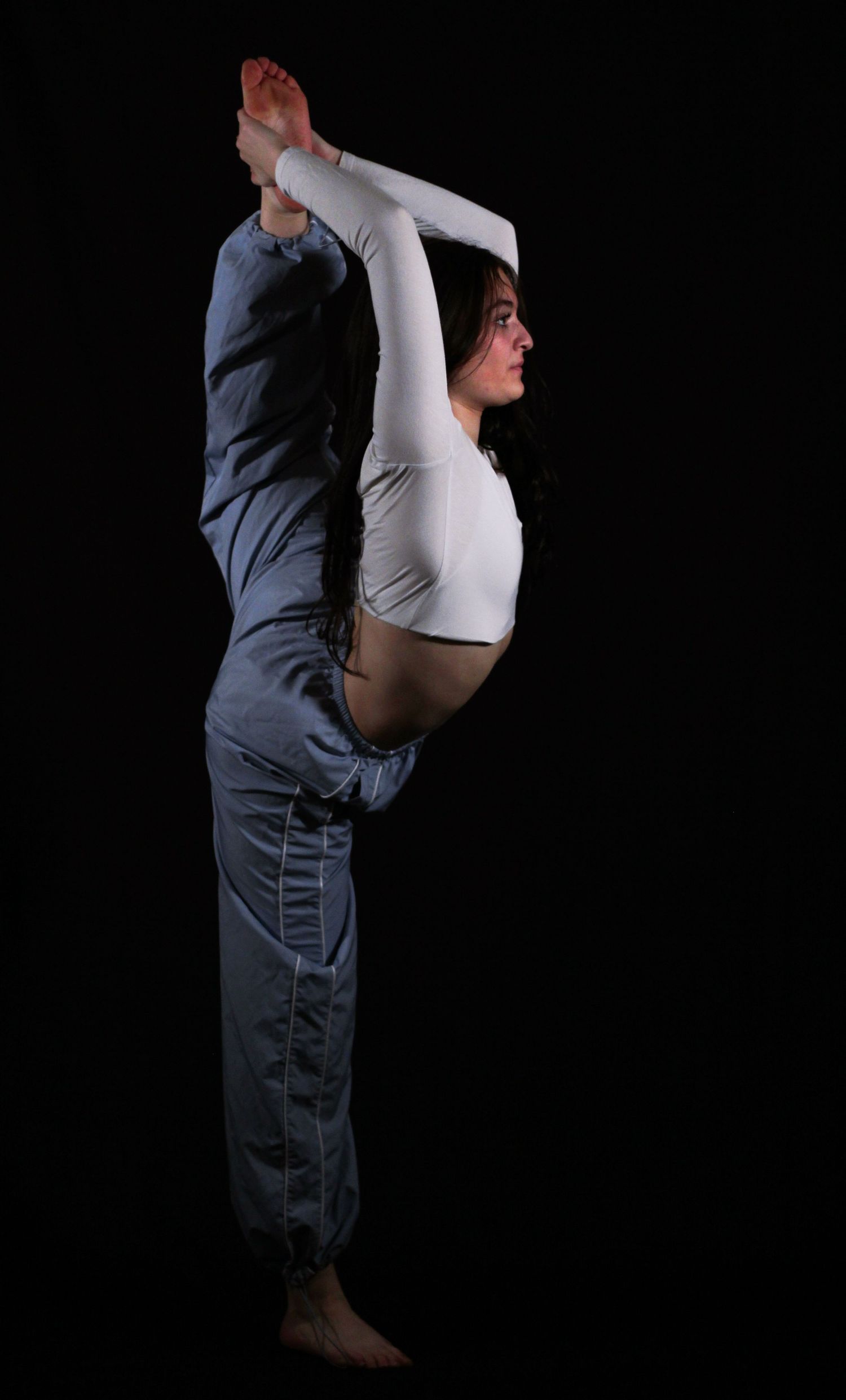 Flexible dancer in a white crop top and gray track pants performs a standing split, holding one leg overhead against a black background.