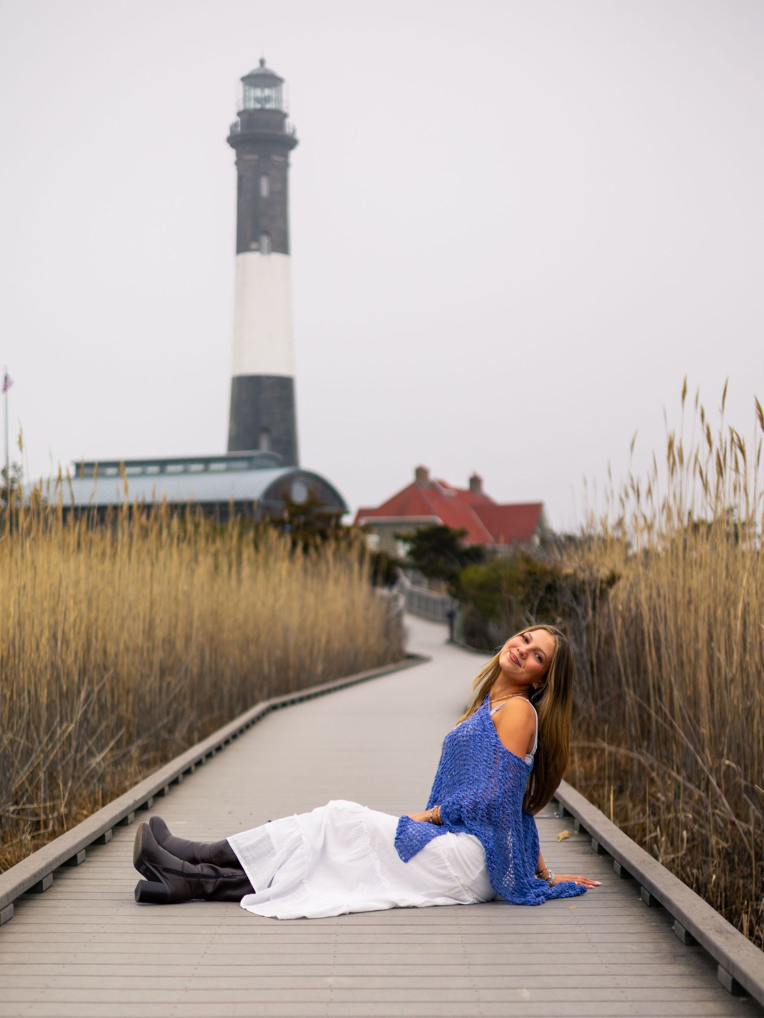 Woman in a blue top and white skirt sits on a boardwalk surrounded by tall reeds, with a black and white striped lighthouse behind her.