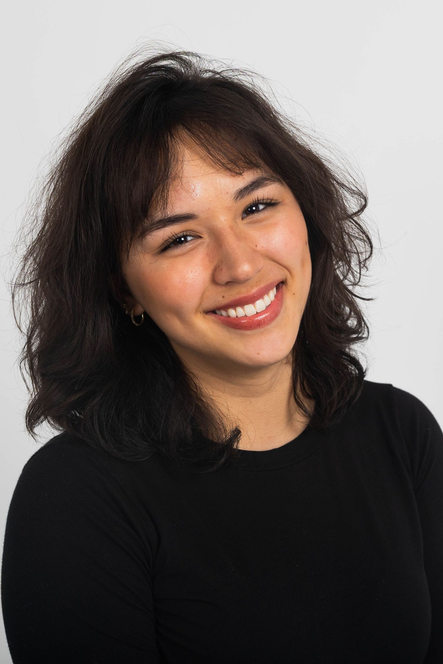 Smiling young woman with shoulder-length dark wavy hair and bangs wearing a black long-sleeve top against a plain light background.