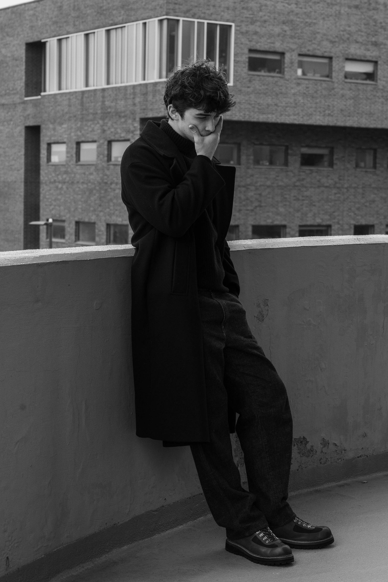 Black and white photo of a young man in a long dark coat leaning on a concrete rooftop wall, hand covering his smile, brick building behind.