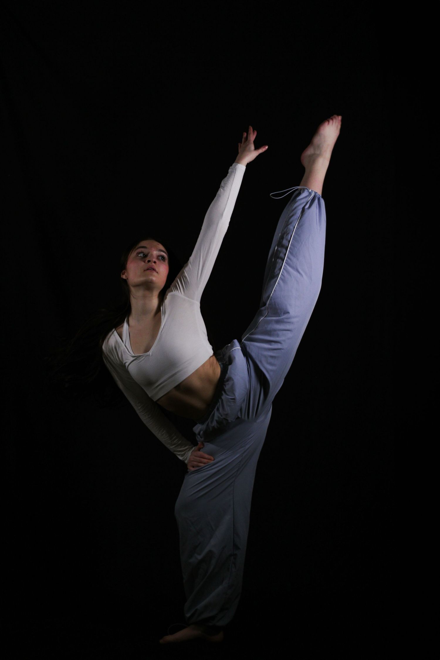 Dancer in a white crop top and light blue pants performs a high standing split with one arm extended against a dark background.