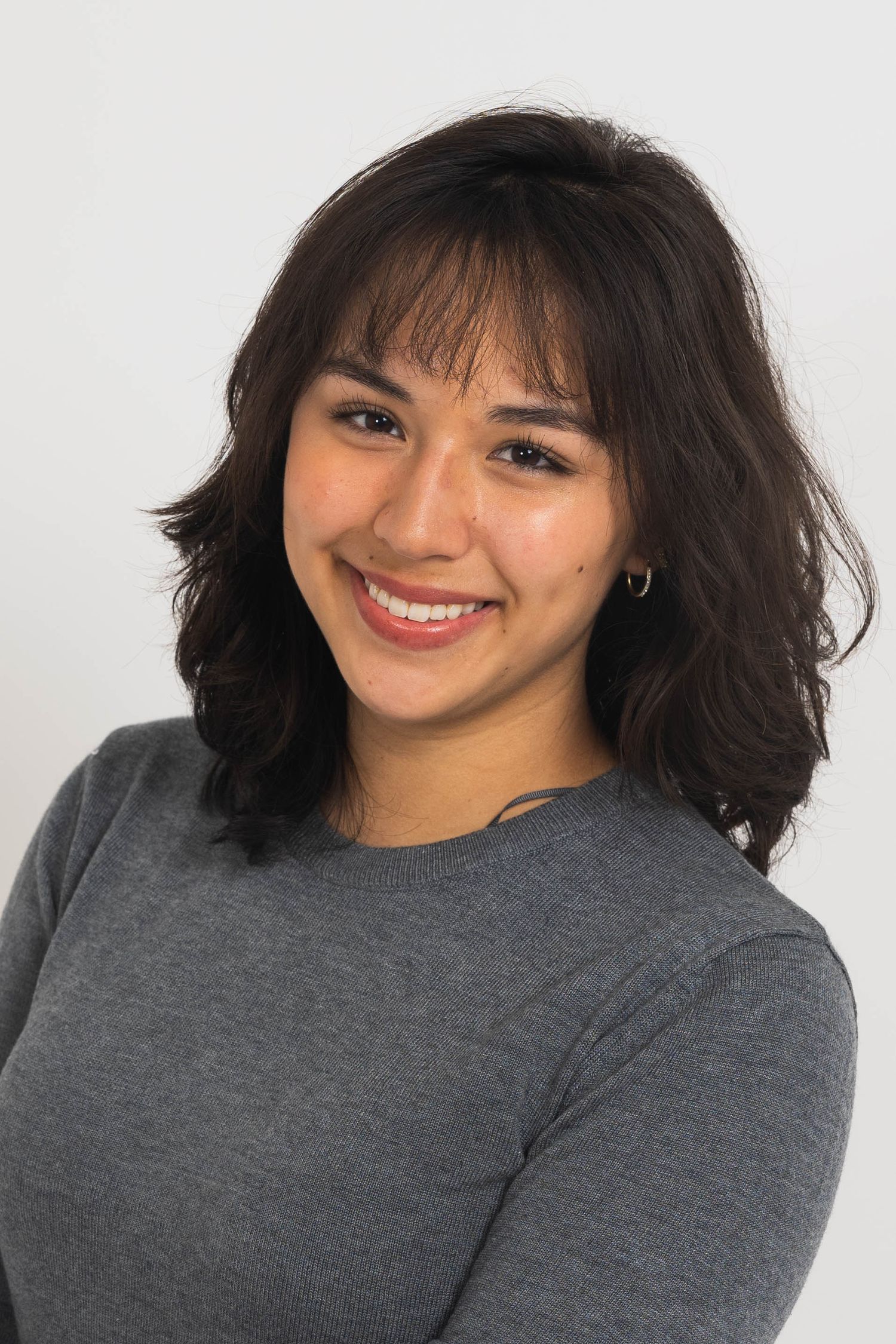 Smiling young woman with shoulder-length dark wavy hair and bangs wearing a gray sweater, posed against a plain light background.