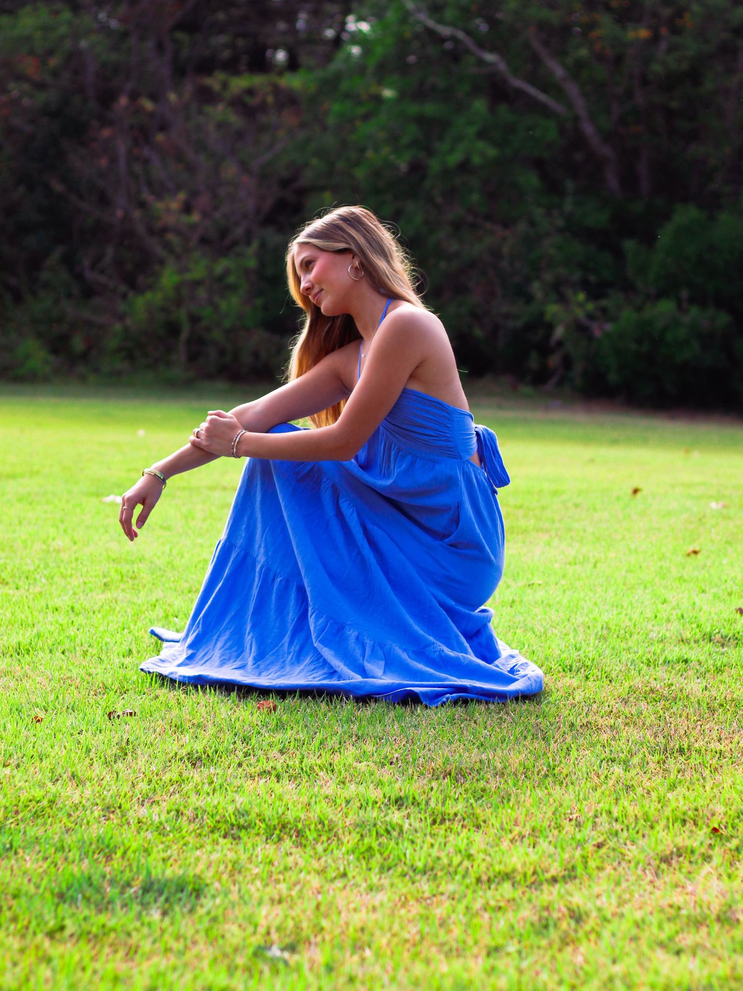 Woman in royal blue strapless maxi dress crouching gracefully on lush green grass outdoors.