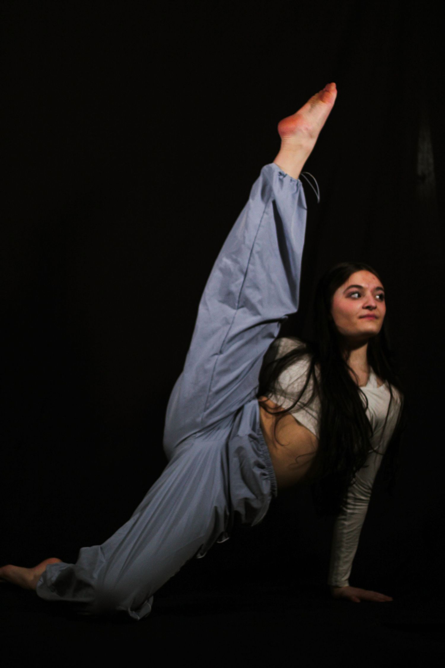 A dancer with long dark hair in loose blue pants performs a high standing split, one leg raised, against a black studio background.