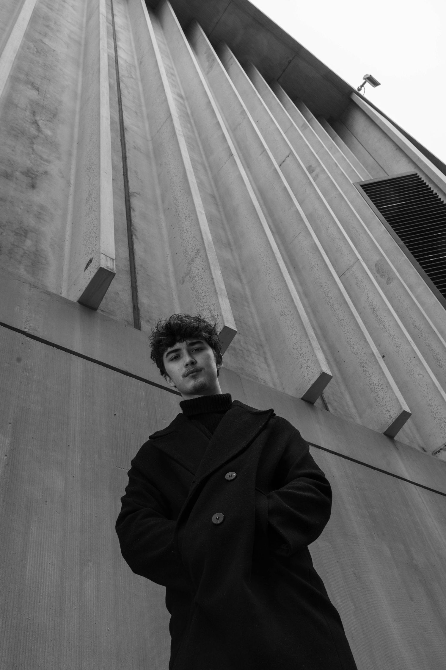 Black and white low-angle portrait of a young man in a dark wool coat and turtleneck standing before a ribbed concrete building.