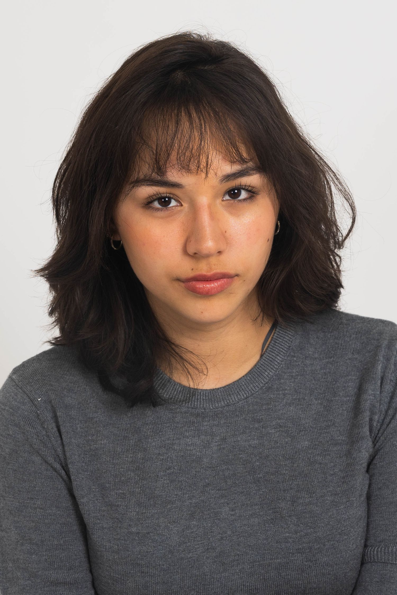 Portrait of a young woman with shoulder-length dark wavy hair and bangs, wearing a gray crewneck sweater against a plain light background.