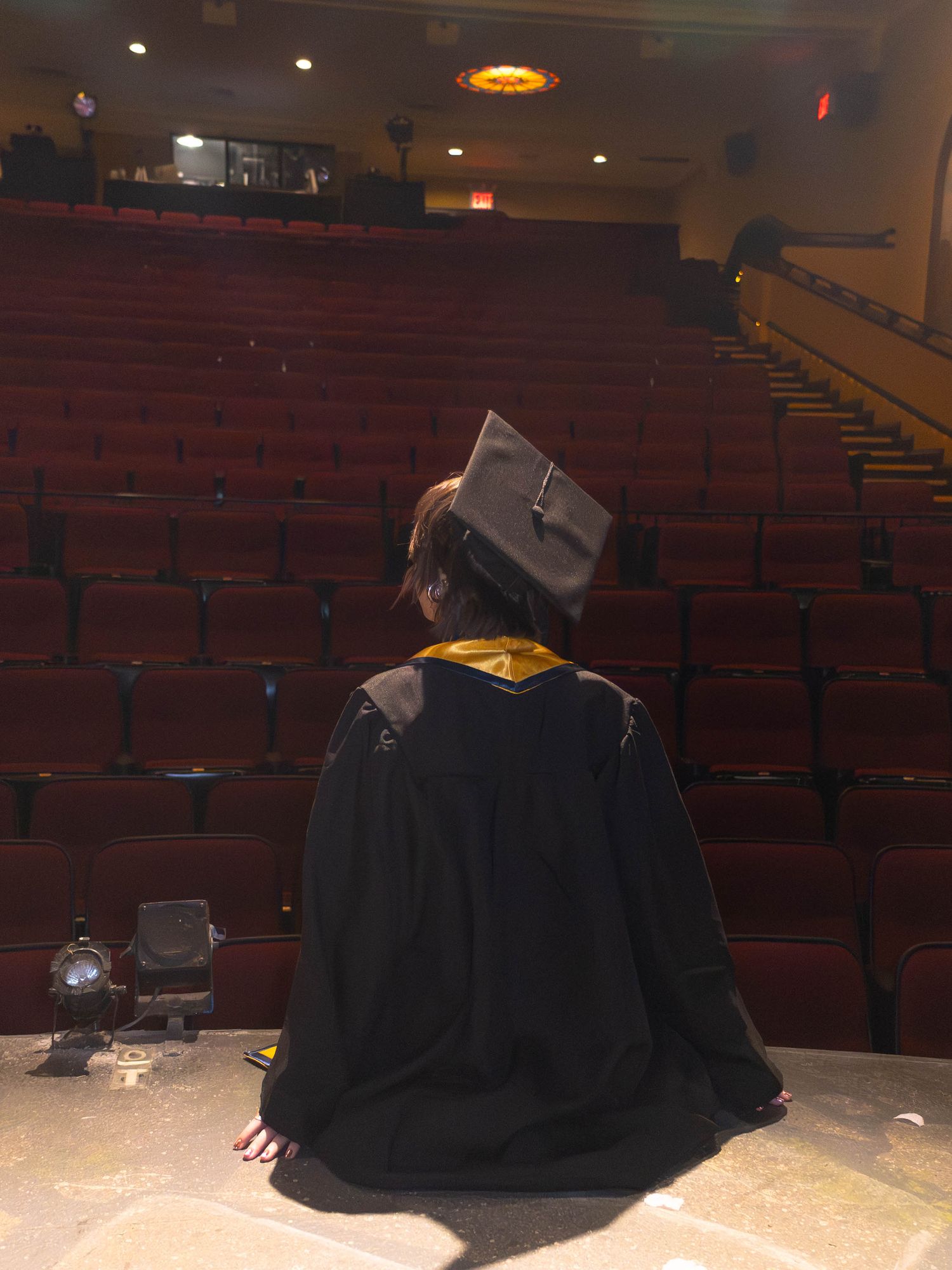 Graduate in cap and gown sits alone on stage facing empty red theater seats before graduation ceremony.
