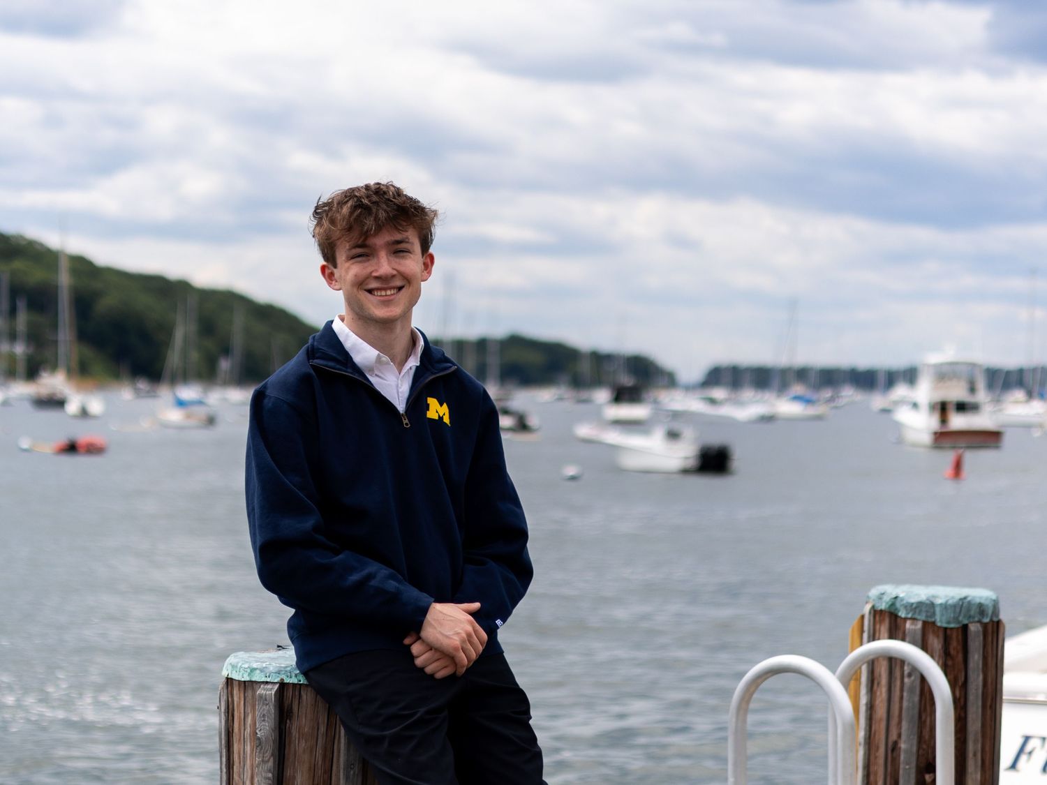 Young man in University of Michigan hoodie smiling on a dock with boats and harbor in the background.