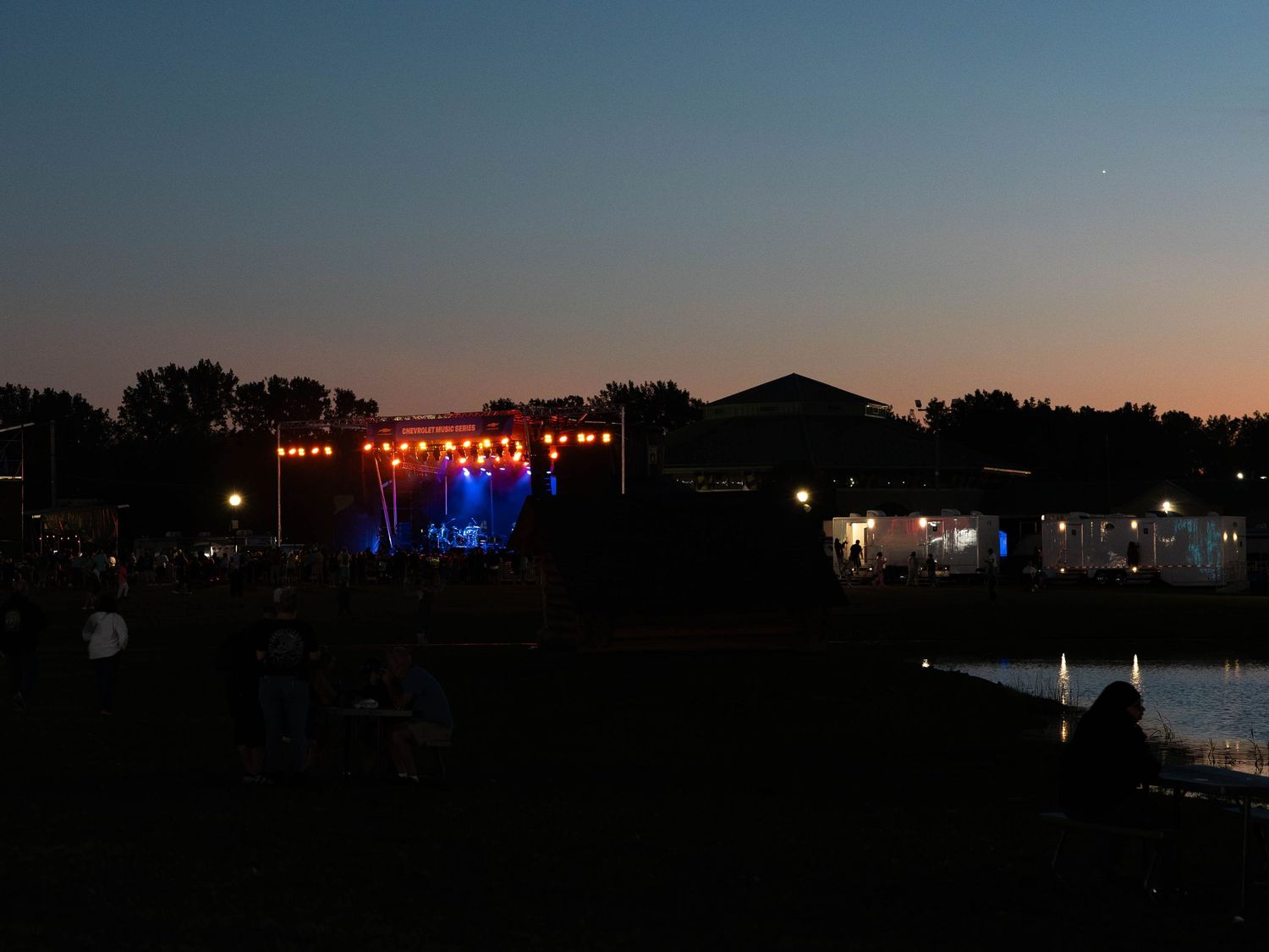 Outdoor concert stage with colorful lights at dusk, crowd silhouettes visible near a pond at a summer music festival.