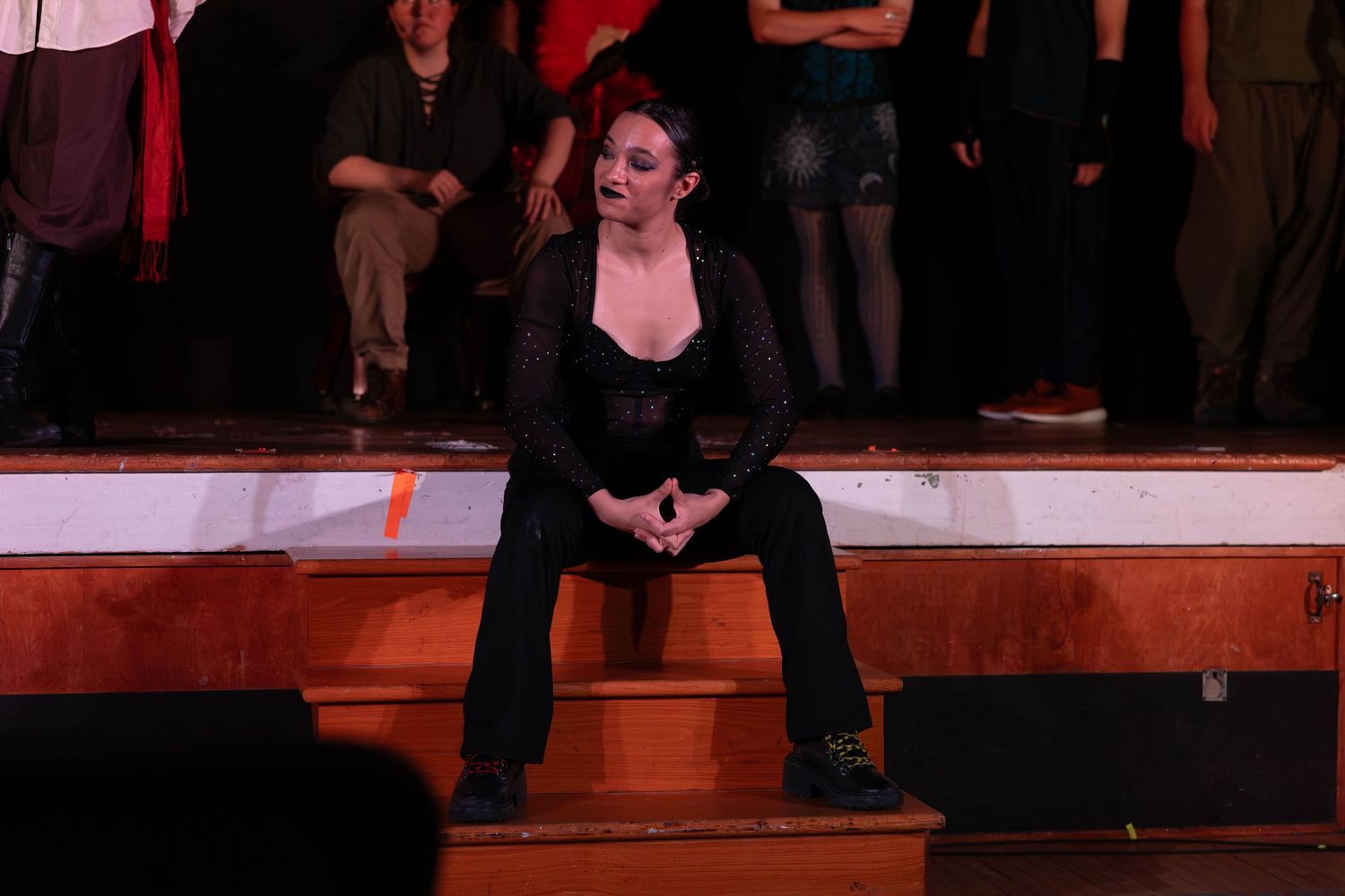 Woman in black outfit sits on wooden stage steps during a theatrical performance.